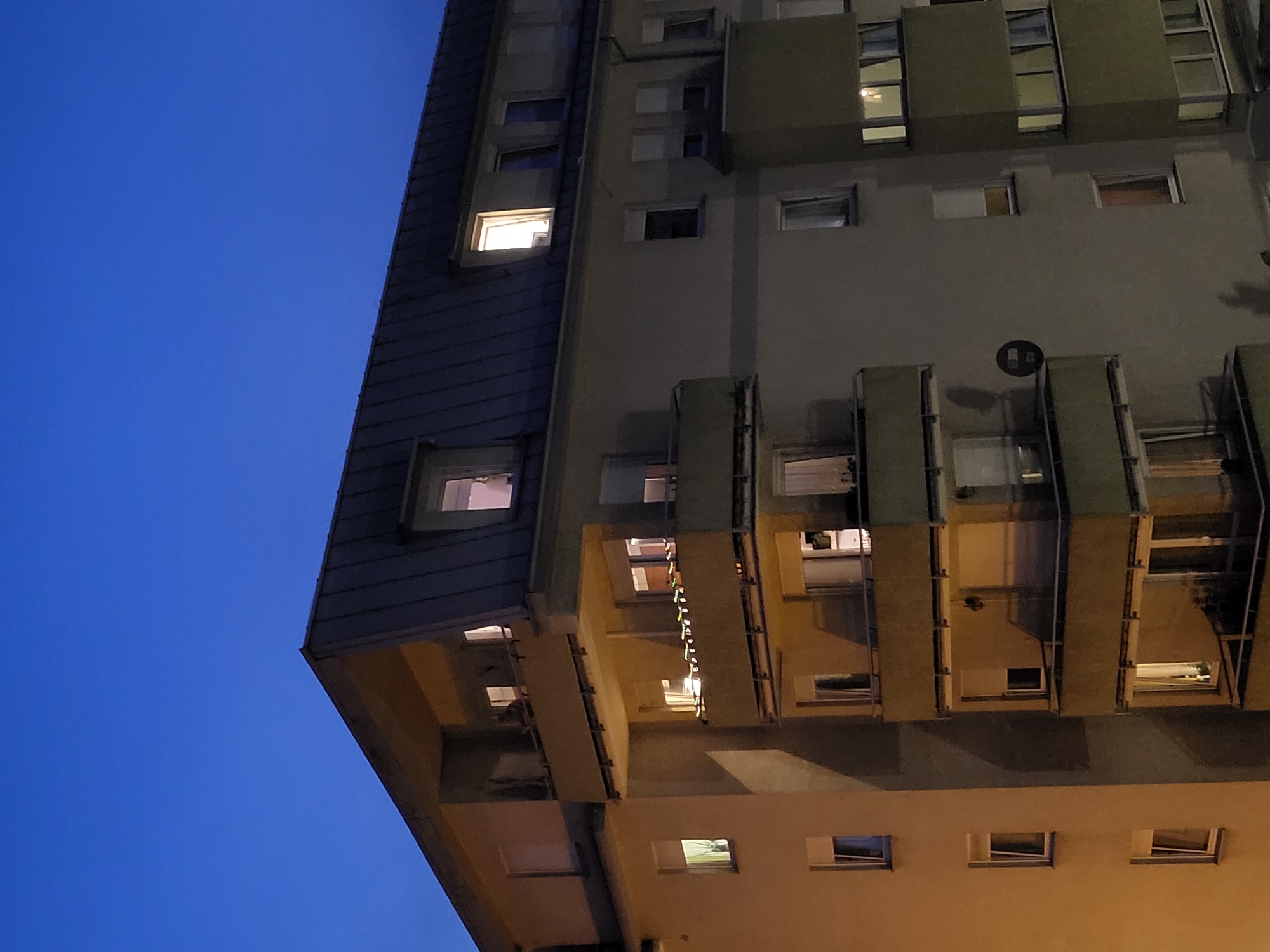 A multi-story apartment building with a dark, slanted roof is shown against a deep blue twilight sky. Several windows are illuminated, suggesting occupancy within the building. Balconies with yellow railings are visible on various levels. On one balcony, a string of small lights is draped along the railing. A circular, dark object, possibly a satellite dish, is mounted on the building's facade. The building appears to be situated in an urban environment.