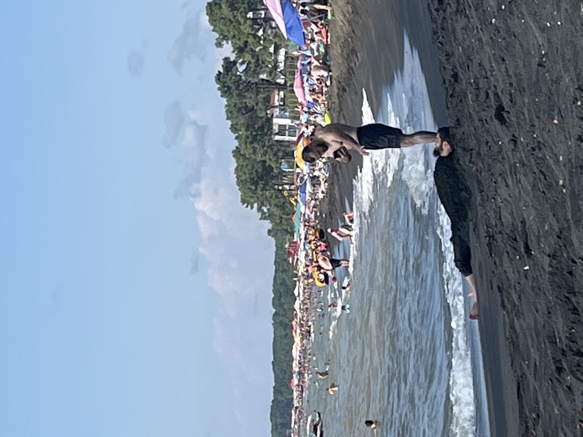 The image depicts a crowded beach scene in Ureki, Georgia. The beach has black sand. In the foreground, two men are interacting on the shore. One man is lying face down on the sand with his head towards the water, wearing dark clothing. Another man, wearing black shorts, is standing over him, looking at his phone. In the background, the ocean is filled with people swimming and playing in the water. Numerous beach umbrellas are set up on the sand, with more people relaxing under them. A line of trees and some buildings are visible behind the beach. The sky is blue with some scattered clouds.