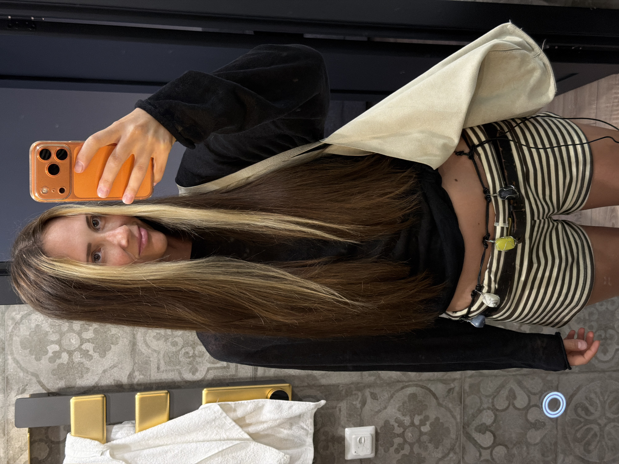 The image shows a woman taking a mirror selfie in a bathroom. She is wearing a black long-sleeved top, striped shorts, and a decorative belt. She has long, brown hair with blonde highlights and is holding an orange-cased smartphone. A beige bag is slung over her shoulder. The bathroom features patterned tiles on the walls, a gold and gray towel rack with a white towel, and a light switch. A blue circular light is visible on the wall. There are no identifying features or landmarks indicating the location is Saratov, Russia.