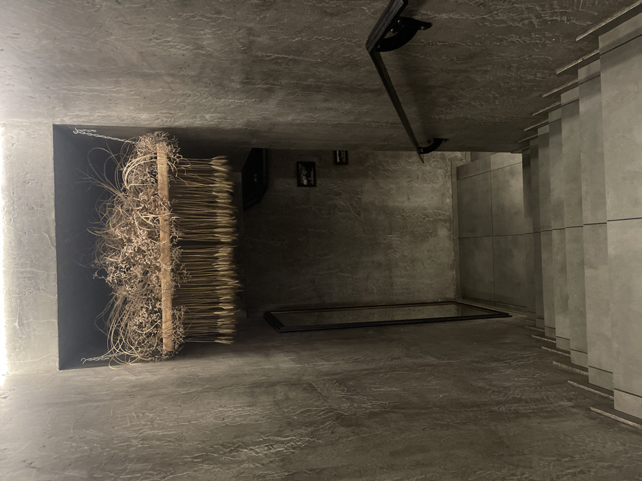 A dimly lit room with a concrete floor and ceiling. On the left, a black wall displays a rustic art installation featuring dried plants and wheat. In the center, a rectangular frame lies on the floor. A black metal bar with mounts is suspended from the ceiling. A stack of vertical grey panels lines the right wall. Two small framed pictures are visible further down on the grey wall.
