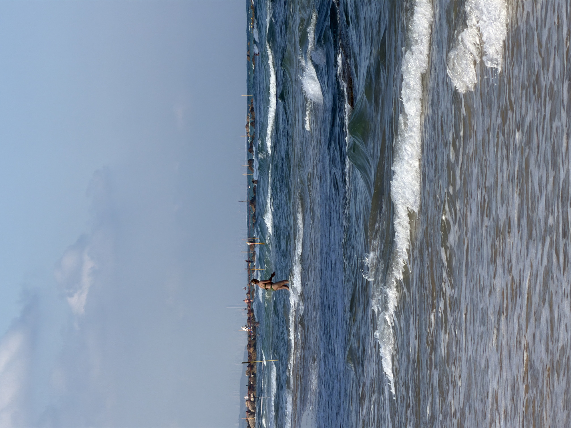 The image depicts a view of the Mediterranean Sea from the coast of Bat Yam, Israel, on a bright, partially cloudy day. The foreground is dominated by the ocean, with waves actively breaking and foaming. In the mid-ground, a woman in a bikini is wading through the shallow water. Behind her, a line of dark, rocky breakwaters stretches across the sea, punctuated by vertical poles. Further in the distance, the silhouette of the Bat Yam skyline is visible, with several buildings rising against the horizon. The sky above is a pale blue, with scattered white clouds.
