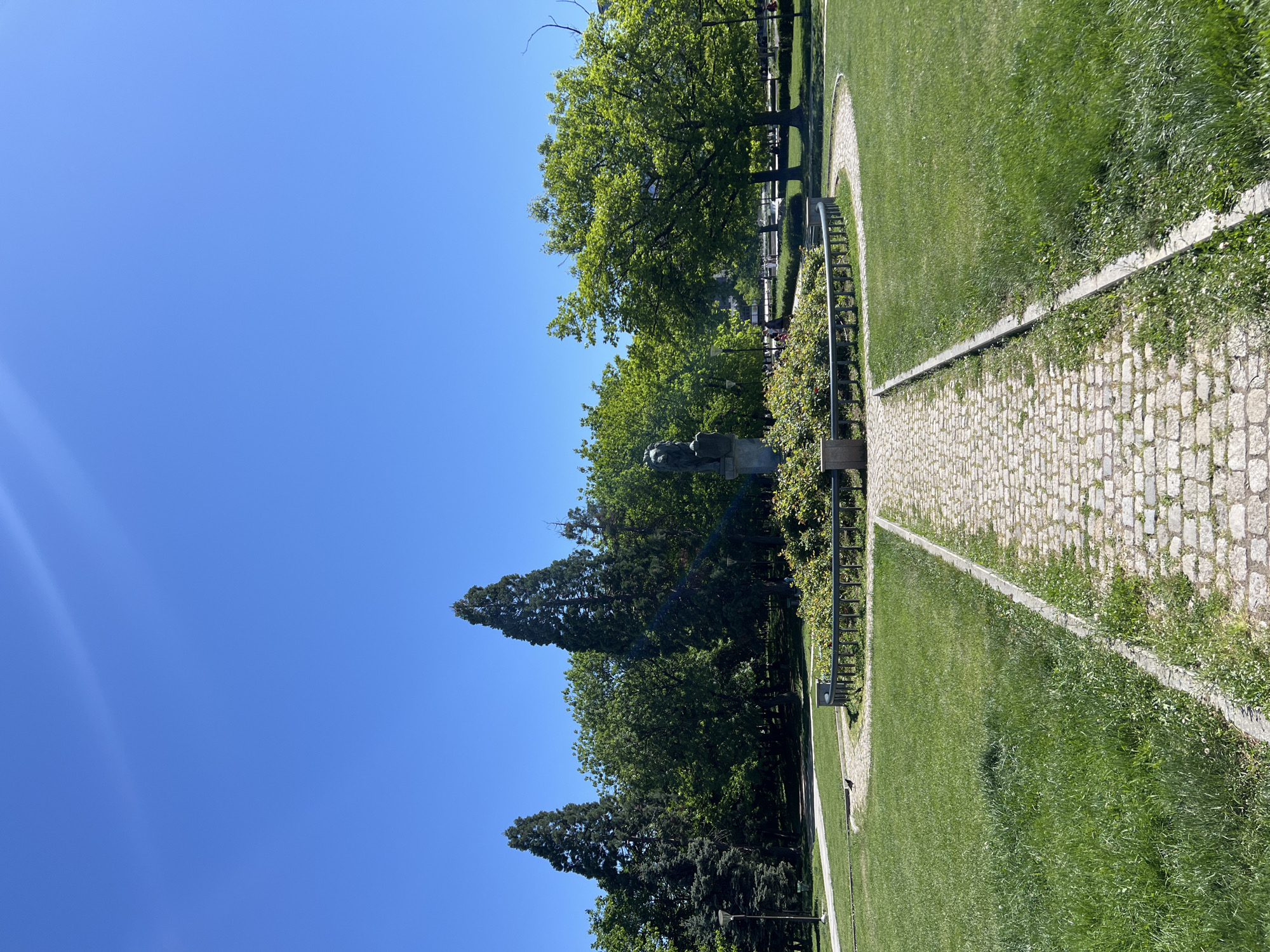 The image captures a park scene in Sofia, Bulgaria. A stone path bisects a green lawn, leading towards trees and a statue. Benches line the path. The sky is clear and blue.