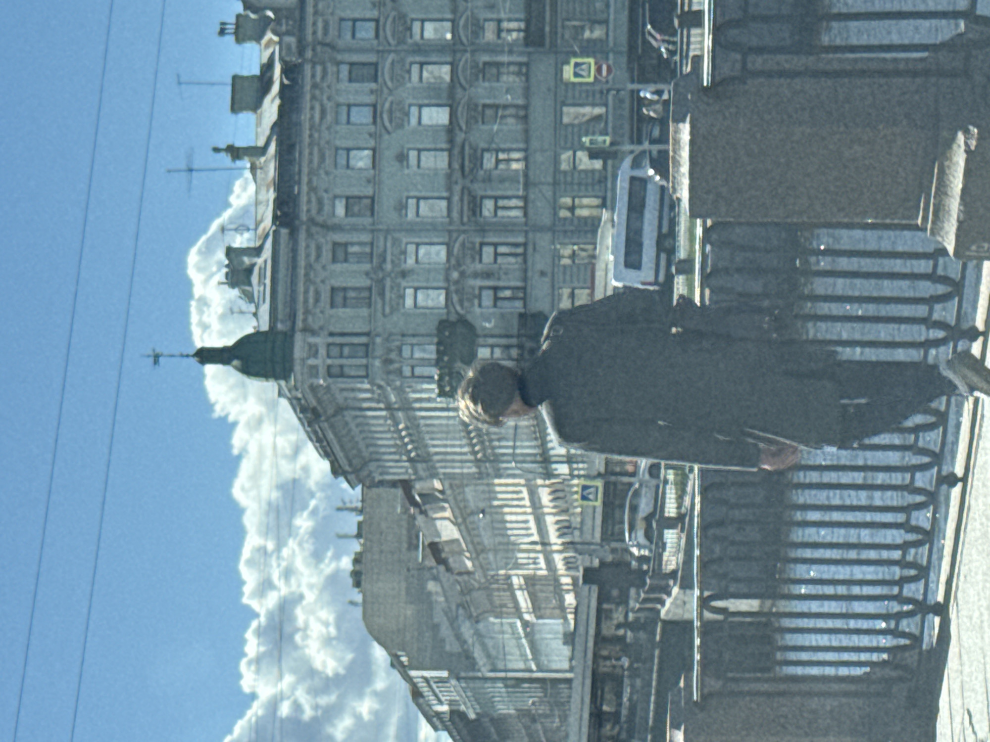The image depicts a street scene in Saint Petersburg, Russia. A person is standing on a bridge next to a fence. In the background, there is an old building, a bus, a sky with clouds, and overhead wires.