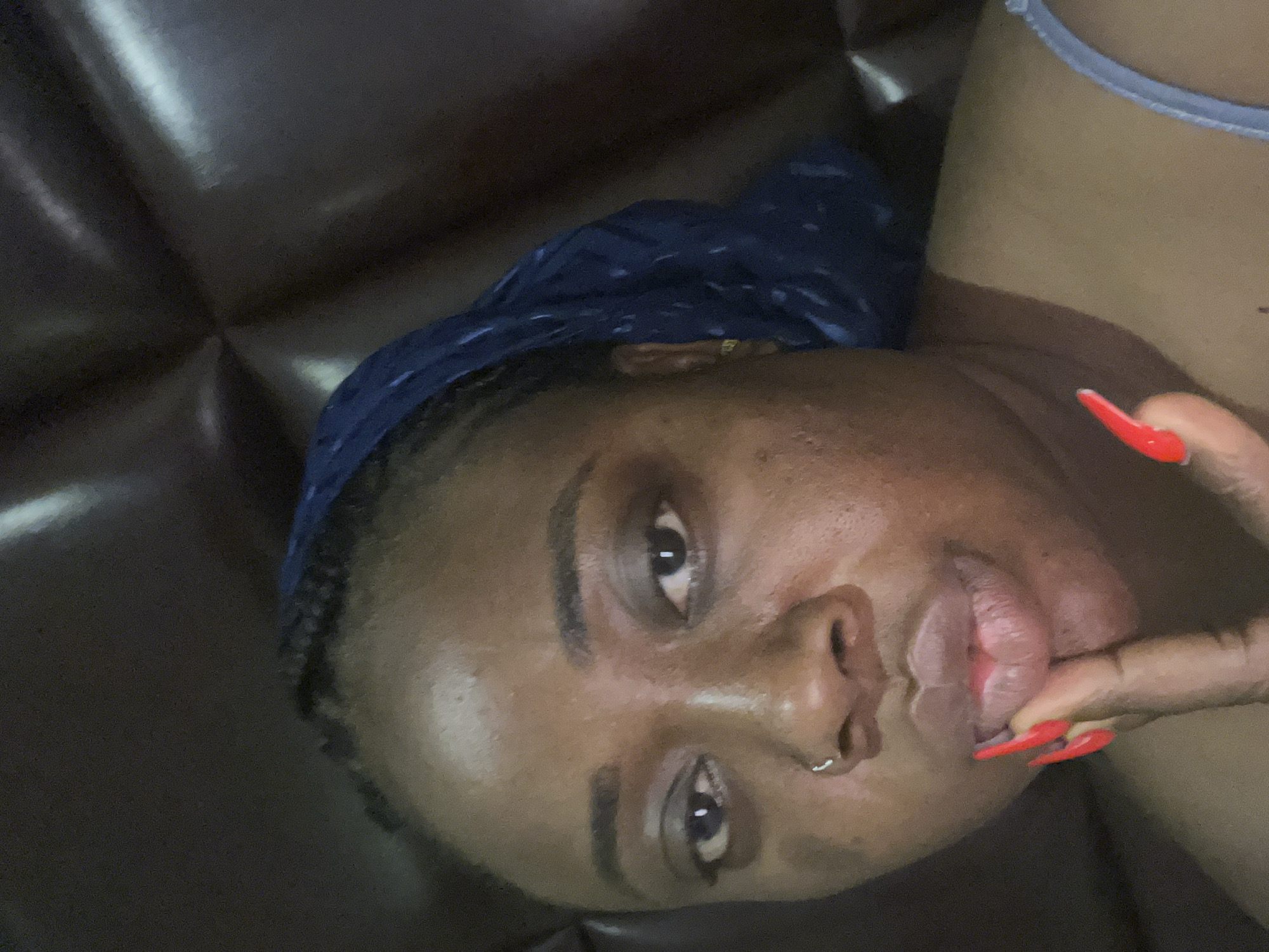 A close-up, rotated portrait features a young person with dark skin, gazing directly at the viewer with a confident expression. Their face is well-lit, highlighting dark eyes, defined eyebrows, and a silver nose ring on their right nostril. A dark blue head covering or bonnet with lighter blue patterns covers their hair, with some braids visible along the hairline.
The person's right hand is raised, with a brightly red-painted finger gently touching their slightly parted lower lip. Their nails are long and sharply shaped. A light-colored strap, possibly from a top or undergarment, is visible on the upper right side of the frame.
The background consists of a dark, textured surface, likely a piece of upholstered furniture such as a leather sofa, showing subtle reflections and creases. The overall setting appears to be indoors, possibly a home in Abuja, Nigeria. The lighting is somewhat soft, creating a focused and intimate atmosphere. No visible text is present in the image.