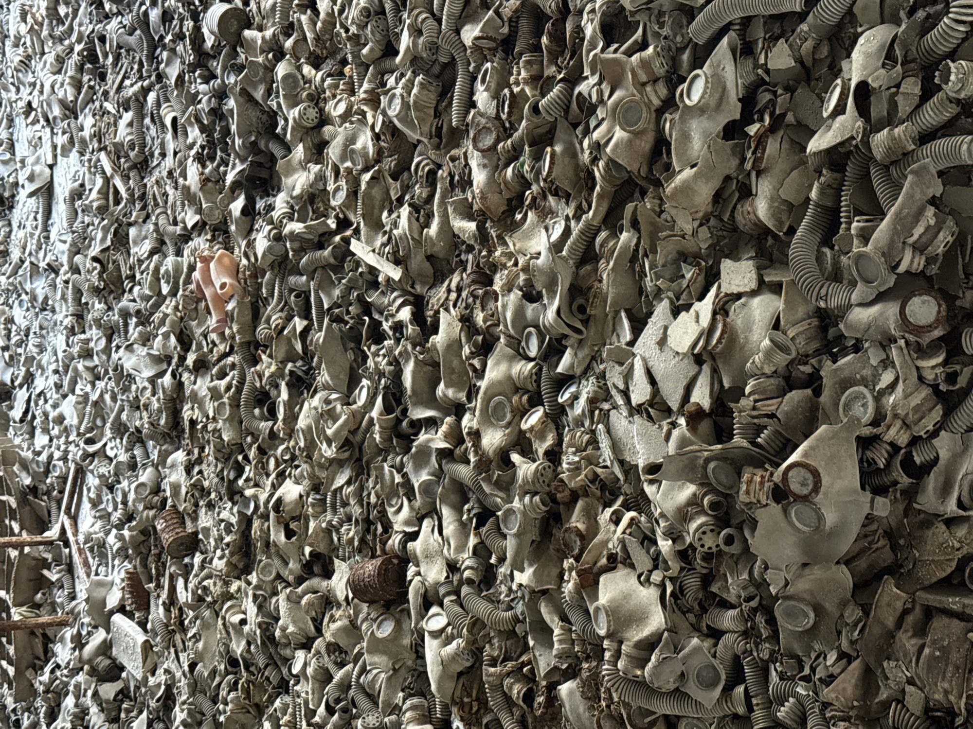 This image shows a wall constructed of discarded gas masks in Prypiat, Ukraine. The masks are mostly gray, with some showing damage or disintegration. Interspersed among the masks are corrugated hoses and filter canisters. On the left of the image, there's a ladder. A pink doll's arm is visible, tucked in between masks.