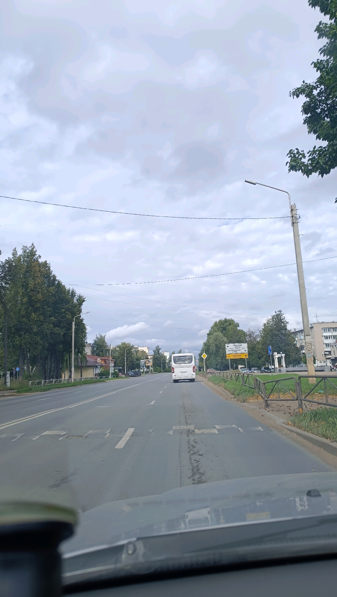 The image shows a view from inside a vehicle looking forward on a two-lane road in Chaikovsky, Russia. The road has lane markings and speed bumps. On the left side of the road, there are trees and some buildings. On the right side, there is a metal fence and grass. Further down the road, a white bus is visible, along with streetlights and buildings. The sky is overcast. There are no people clearly visible in the image, and no specific actions or interactions are depicted. The overall scene is that of a typical urban street on a cloudy day.