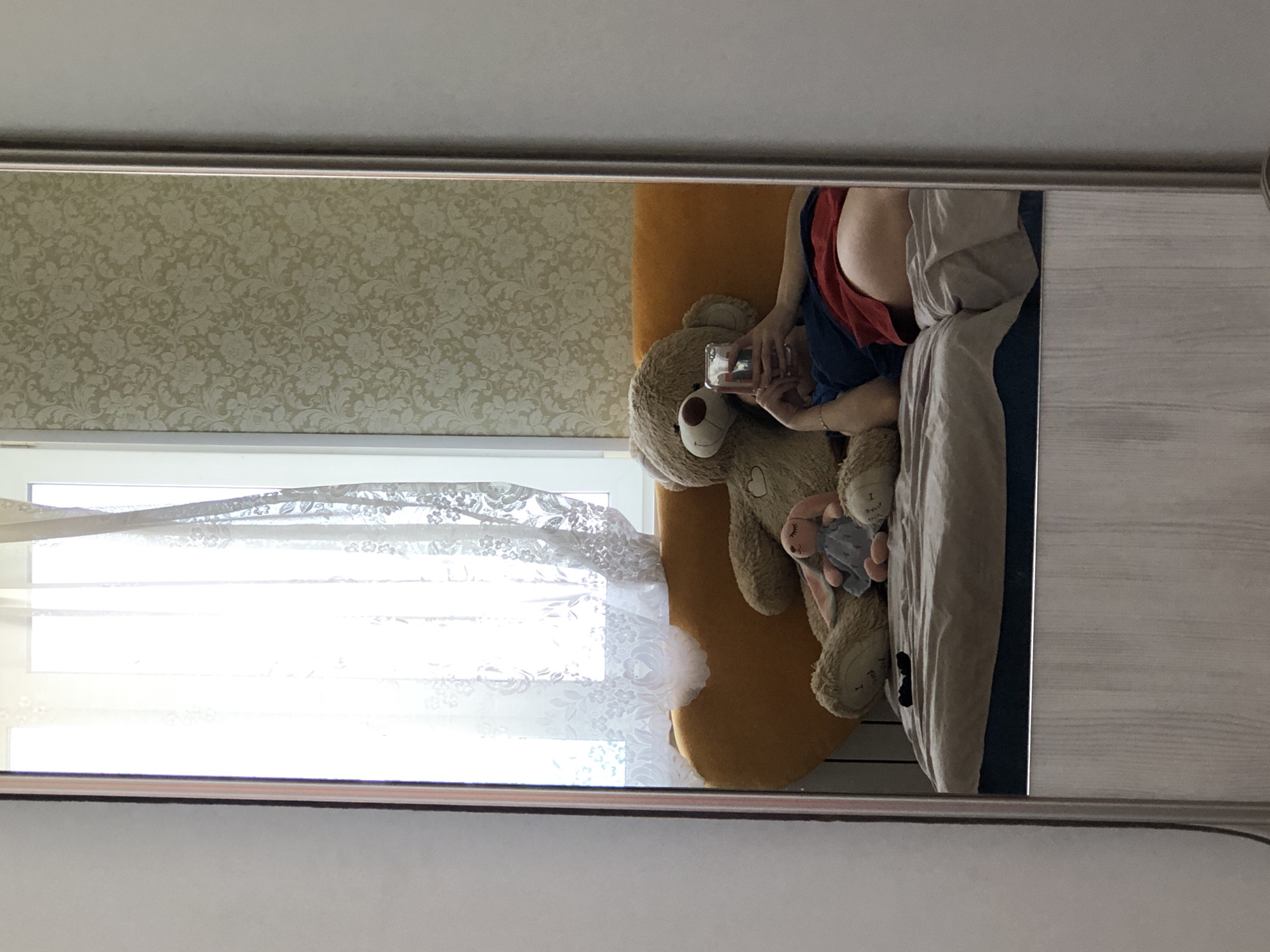 Reflection in a mirror shows a room interior. A person in a red shirt and blue clothing is lying on a bed with a yellow pillow. A teddy bear and other stuffed animals are next to the person. The person is holding a phone. A window with lace curtains is visible. The wall has floral wallpaper. The room is located in Noviy, Russia.