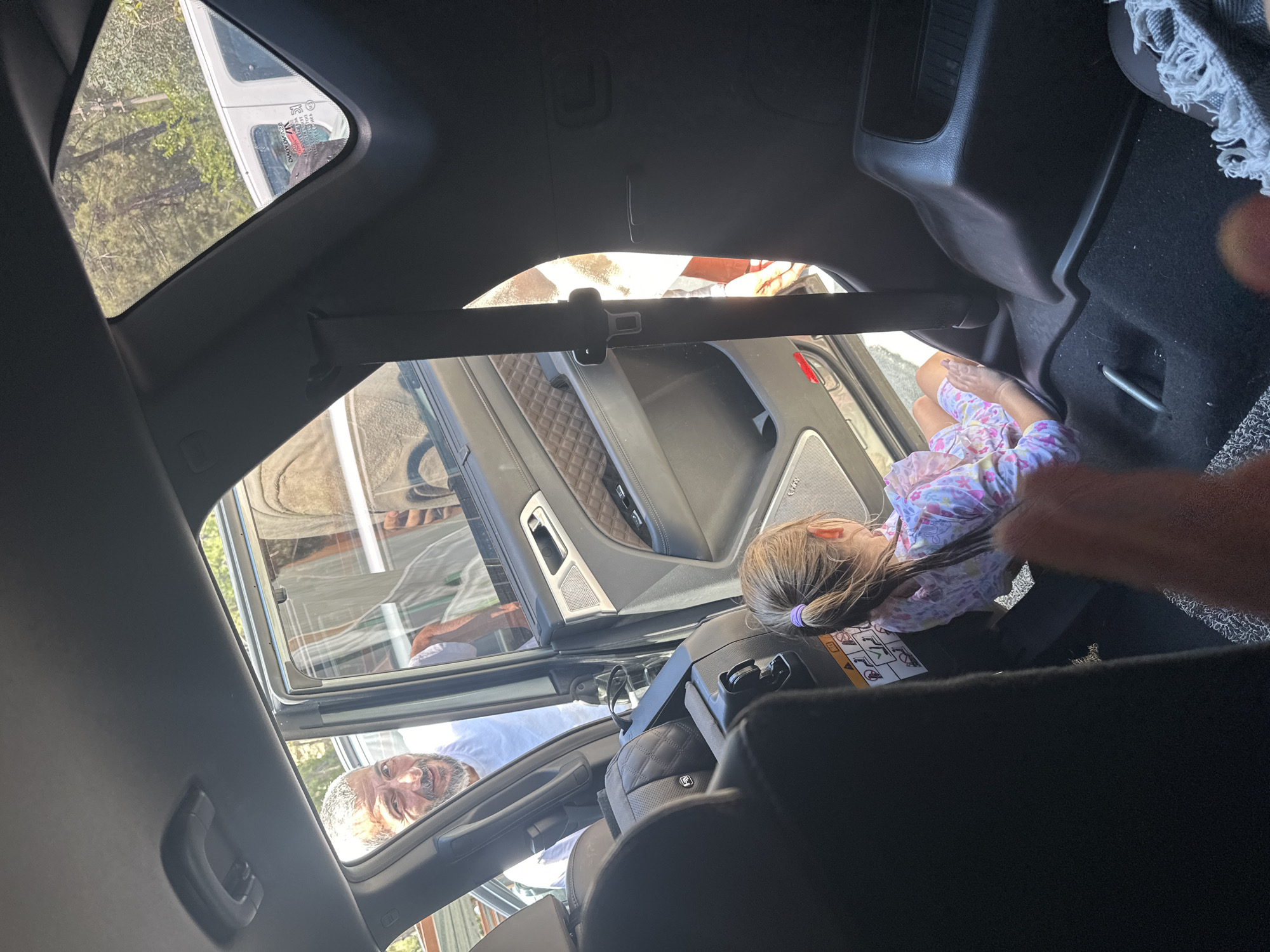 From inside a vehicle, a partial view of a girl in a car seat is seen. Outside the vehicle, a man is visible through the open door. The setting is outdoors, with glimpses of trees and a white van suggesting an urban or suburban environment. Without additional landmarks or information within the image, a specific location in Fethiye, Turkey, cannot be determined.