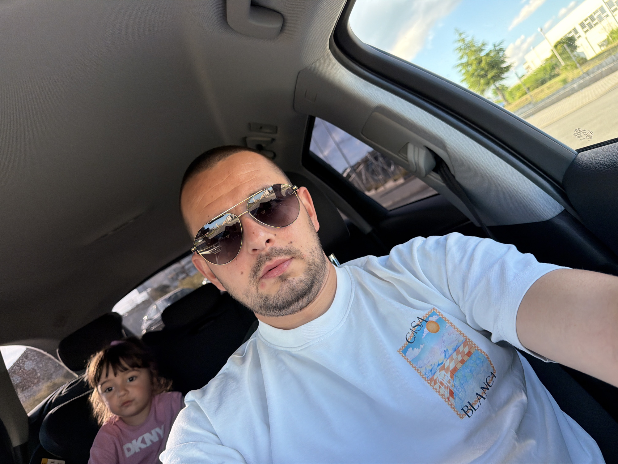 A man with short hair, stubble, and sunglasses is taking a selfie inside a car. He is wearing a white t-shirt with a colorful graphic. In the back seat, a young girl with brown hair and bangs is visible, wearing a pink t-shirt. The car's interior, including the sunroof and parts of the dashboard, is visible. Outside the car, through the sunroof and windows, a bright sky with clouds and trees can be seen, suggesting an outdoor, daytime setting. The scene appears to be on a street in Burgas, Bulgaria, based on the context provided.