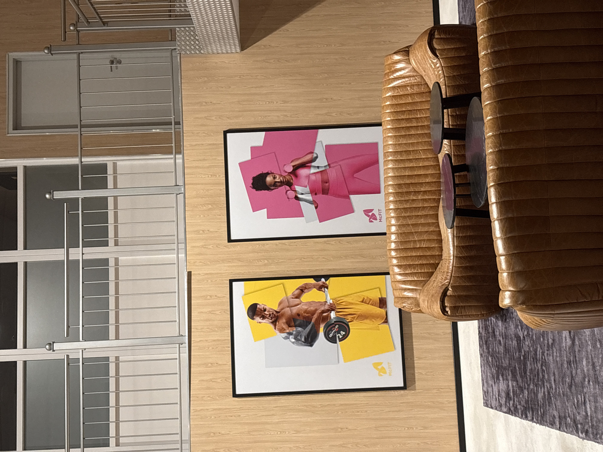This image shows part of the interior of a building, likely a fitness center in Mönchengladbach, Germany. The scene includes two framed posters hanging on a light-colored wooden wall. The top poster features a woman in pink workout attire. The bottom poster features a man lifting weights. In the background, there is a window with a metal security grate. To the right of the posters is a brown leather sofa and a small table with two black circular objects on top. A rug with a gray and white pattern is partially visible next to the sofa.