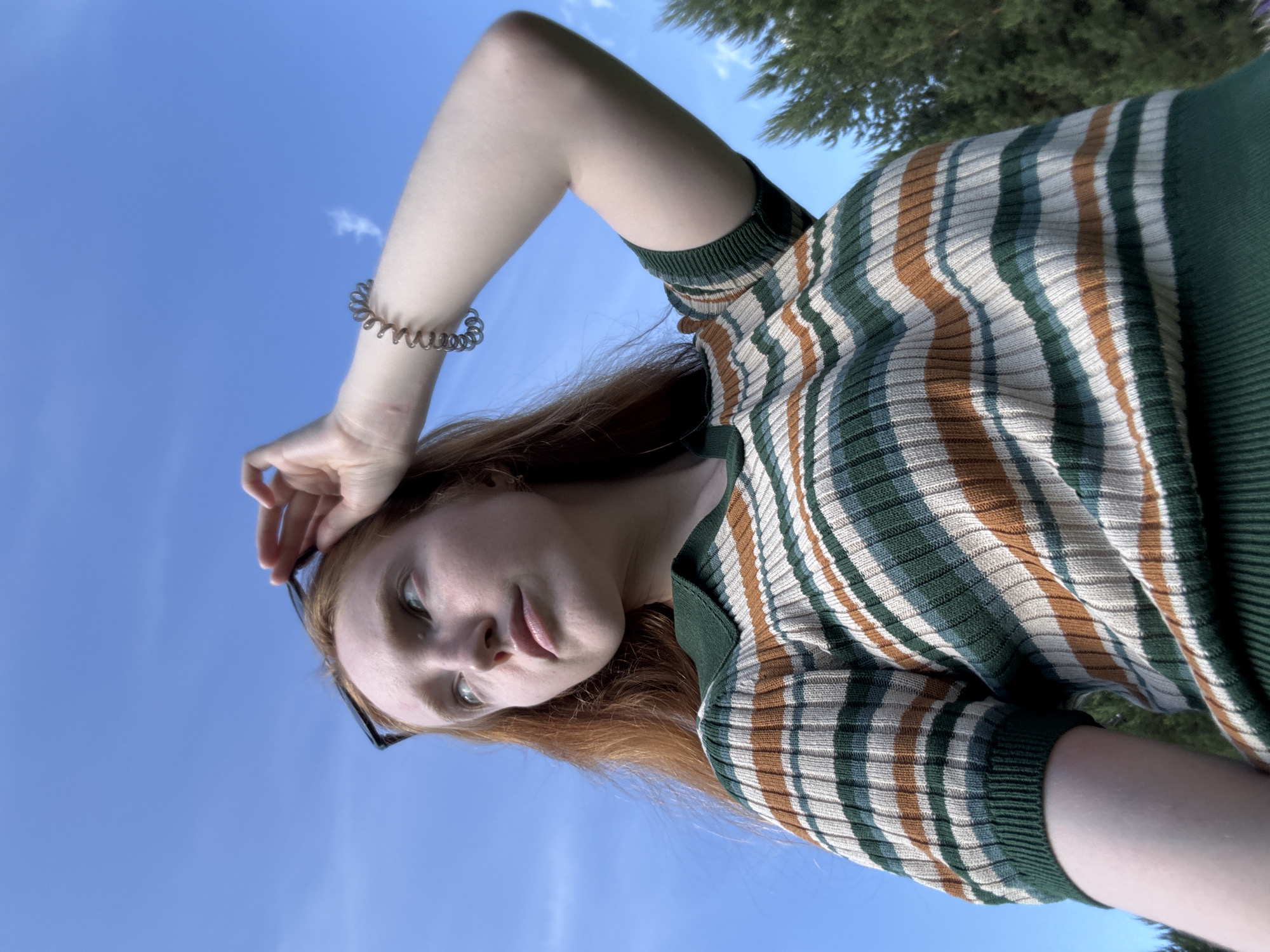 The image displays a fair-skinned young woman with long, reddish-brown hair, viewed from a low-angle perspective, looking upwards towards a bright blue sky with a few wispy white clouds. She is wearing a ribbed knit short-sleeved polo shirt with horizontal stripes in varying widths, featuring colors such as dark green, light green, beige, and rusty orange. The collar and sleeve cuffs are solid dark green. Her right hand is raised to her head, lightly touching a pair of black-framed sunglasses perched on top of her hair. A dark-colored spiral hair tie is visible on her right wrist. In the lower right background, the tops of dark green coniferous trees are partially visible. This scene is set outdoors in the city of Chaikovsky, Russia.