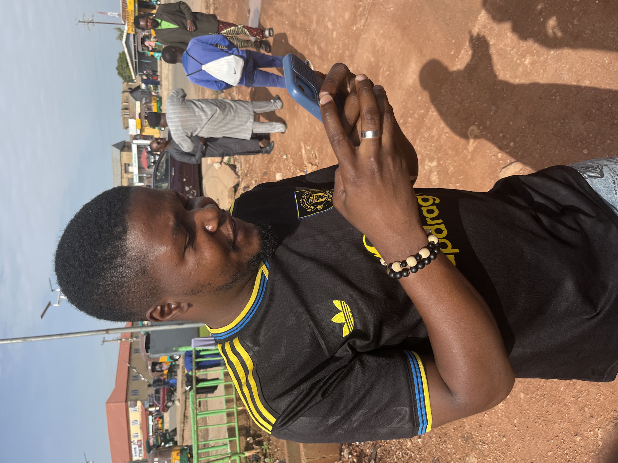 An outdoor, eye-level, medium shot shows a Black man in Gura Top, Nigeria, looking down at his blue smartphone. The man is wearing a black, short-sleeved soccer jersey with yellow and blue stripes on the sleeves and collar. He also has a silver ring on his left ring finger and a bracelet made of black and wooden beads on his right wrist. The man's face is partially obscured by his hands and the phone. He appears to be in a street or market area, with a dusty, reddish-brown ground and several other people in the background. Some of the background figures are blurred, suggesting motion or a shallow depth of field. Buildings and vehicles, including what appear to be auto-rickshaws, are visible in the distance, indicating an urban or semi-urban setting. The lighting suggests it is daytime, with bright sunlight creating shadows on the ground.