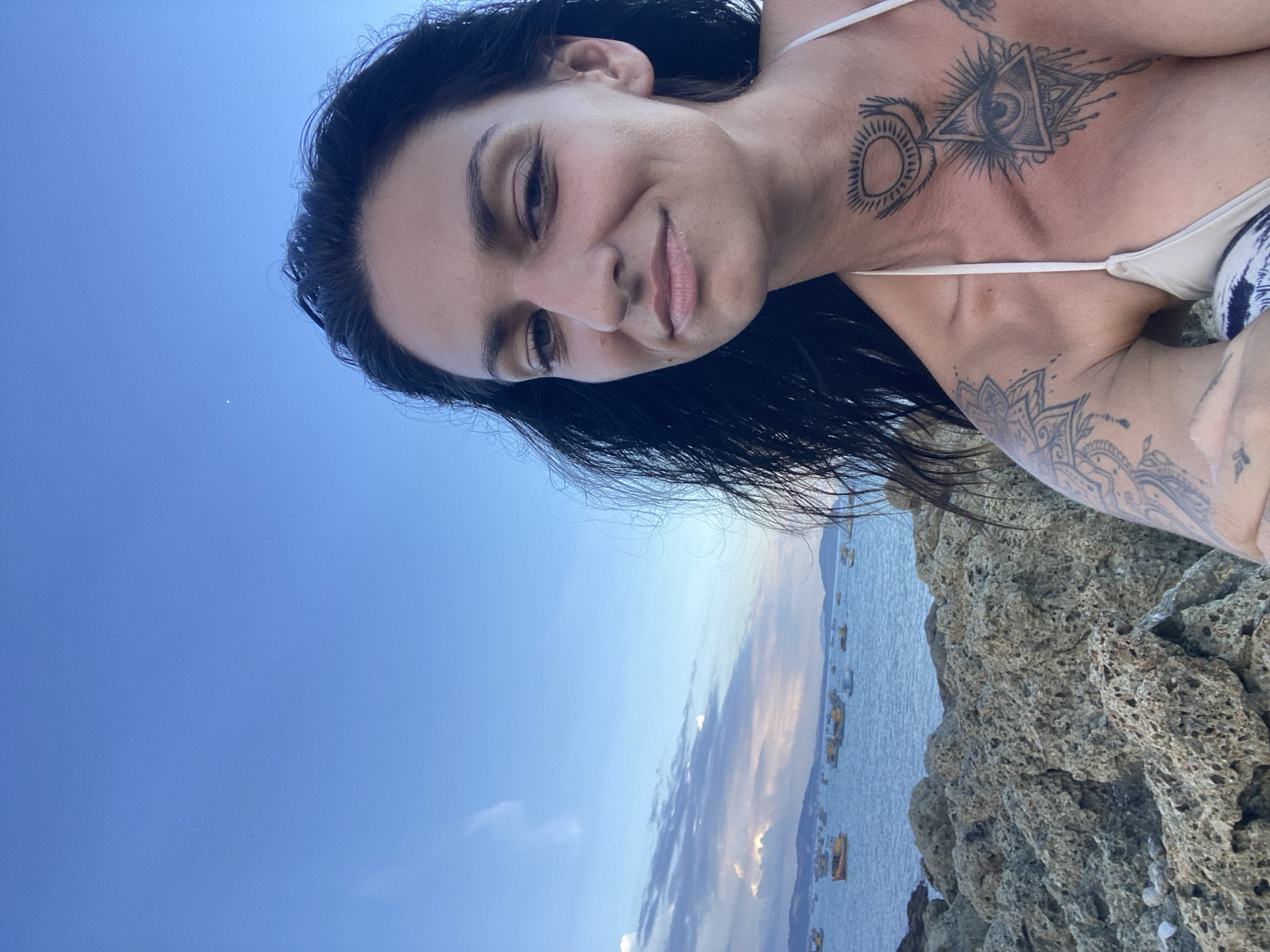A selfie captured on a rocky shore in Mũi Né, Vietnam, shows a woman with long, dark, wind-swept hair and tattoos on her chest and shoulder. She is wearing a bikini top. In the background, the sea is dotted with numerous small fishing boats. The sky above is a clear blue, with a few wispy clouds and a sliver of the moon visible. The foreground is dominated by rough, grey rocks. The overall scene suggests a coastal landscape under a daytime sky.