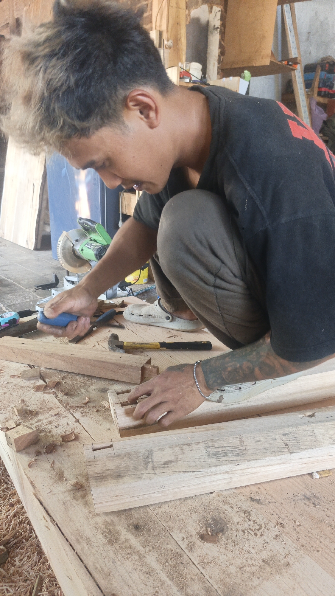 In a dimly lit workshop in Kandangmas, Indonesia, a young man with dyed blonde hair and a black t-shirt is engrossed in woodworking. He is crouched over a workbench, intently focused on joining two pieces of wood. His left hand holds a chisel with a blue handle, poised to shape the wood, while his right hand delicately positions another wooden piece, revealing a dovetail joint. A hammer with a yellow handle rests nearby, as does a miter saw in the background. The floor is scattered with wood shavings, indicating active work. The man's arms are covered in tattoos, and he wears a simple silver bracelet. The overall impression is one of focused craftsmanship amidst a rustic, working environment.