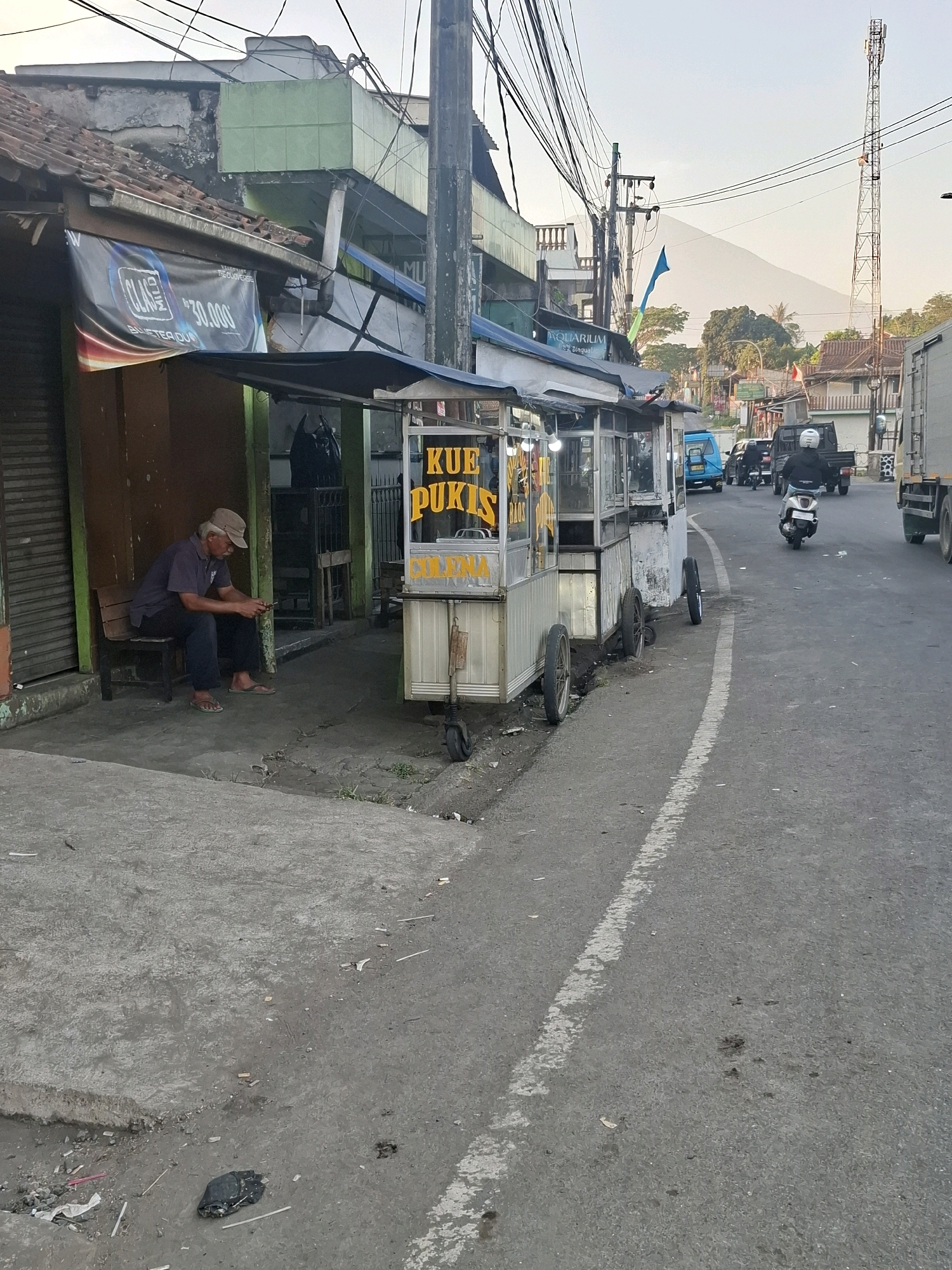 The image depicts a street scene in Sukaraja, Indonesia. In the foreground, a man wearing a cap and casual clothing sits on a wooden bench, looking at his phone. Adjacent to him is a food cart with "KUE PUKIS" written on it in yellow letters, suggesting it sells traditional Indonesian cakes. The cart has wheels and appears to be made of metal. A street runs alongside the sidewalk, with a white dashed line marking the lane. Several vehicles, including a scooter with a rider wearing a helmet, a blue van, and a black truck, are visible on the road. In the background, a cell tower stands tall against a hazy sky. The buildings lining the street show signs of wear, with some having peeling paint. The overall environment suggests a somewhat urban or semi-urban setting.