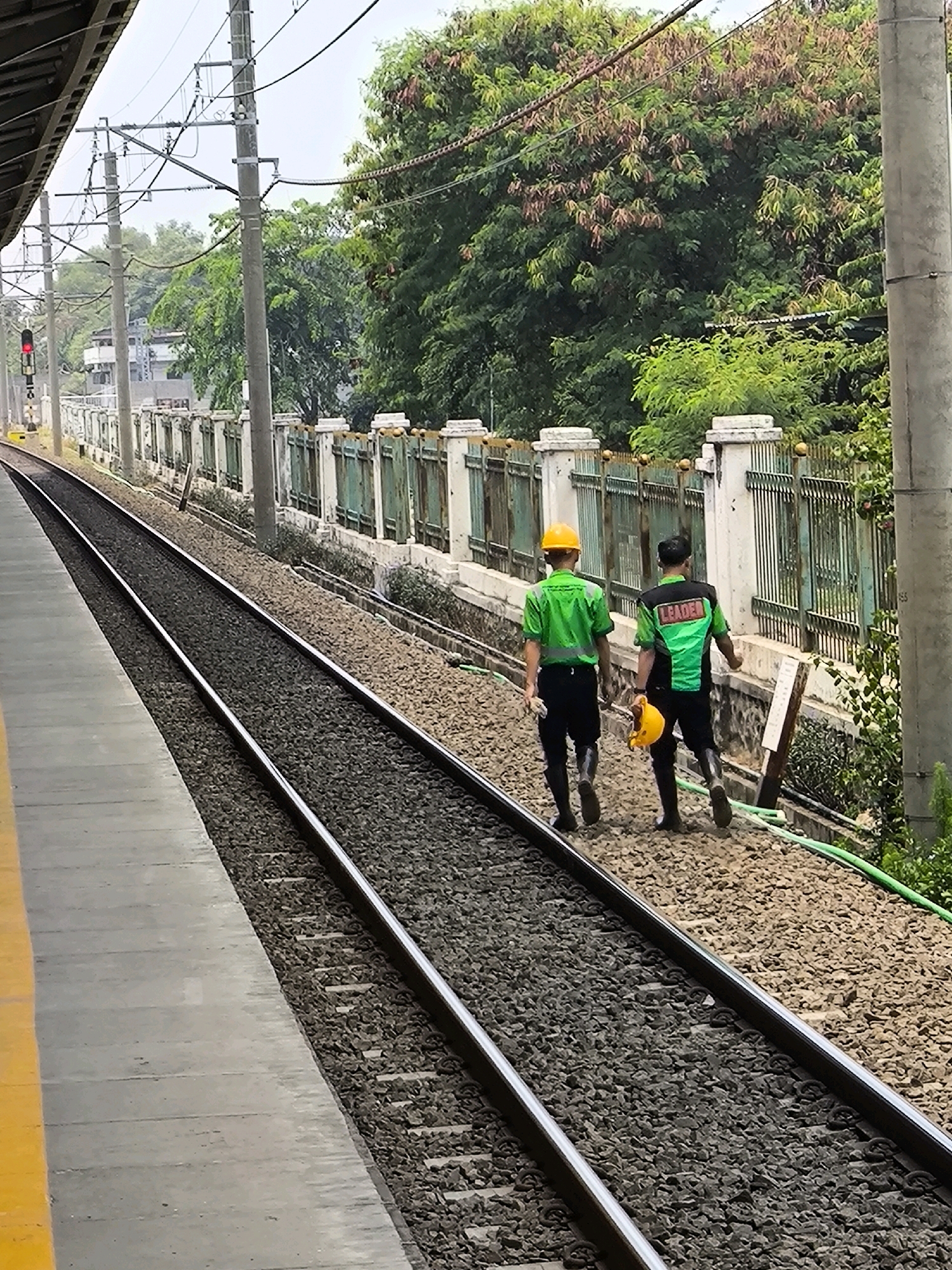 Two men in high-visibility green shirts and hard hats walk along a railway track. The man in front is wearing a yellow hard hat and the man behind him is wearing a green hard hat. The man behind has "LEADER" written on the back of his shirt. They are both wearing black pants and dark boots. The man behind is carrying a yellow hard hat in his left hand. The railway track is made of gravel and steel rails. In the background, there is a metal fence with decorative pillars and dense green foliage. A traffic light is visible in the distance, showing a red signal. The scene appears to be in an outdoor, possibly industrial or transportation-related area.