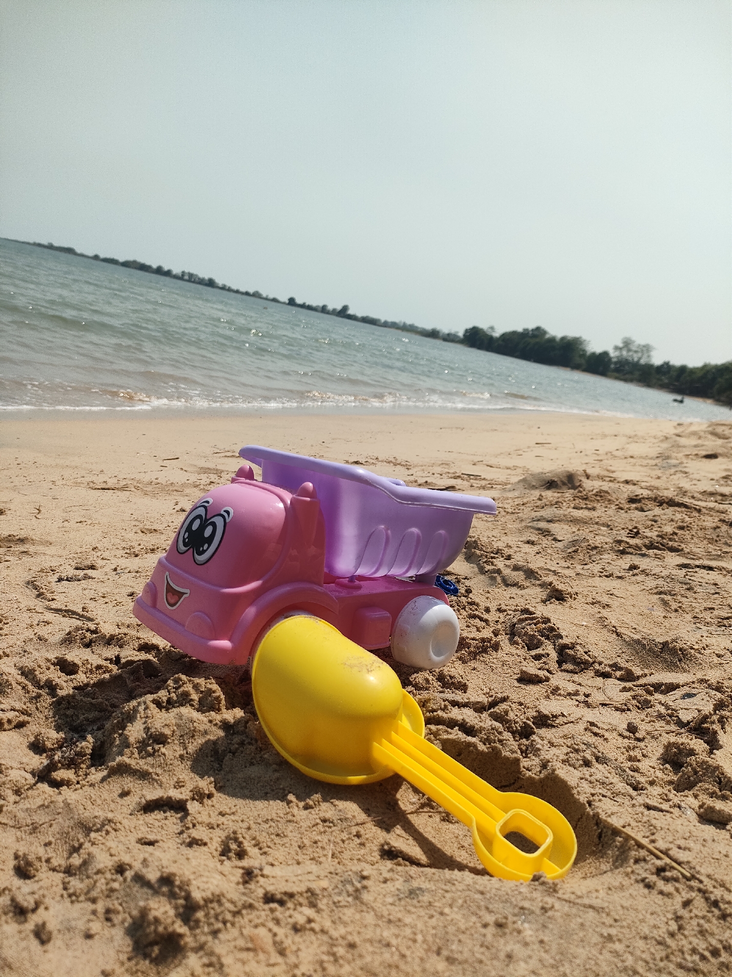 A close-up, low-angle shot captures a pink and purple toy truck and a yellow toy shovel resting on a sandy beach. The truck has a smiling face with large cartoon eyes. Behind the toys, the gentle waves of a body of water lap at the shore, and a distant treeline is visible under a hazy sky. The scene suggests a relaxed, outdoor environment, possibly a park or recreation area. The beach is comprised of fine, light brown sand. The image has a shallow depth of field, with the toys in sharp focus and the background softened.