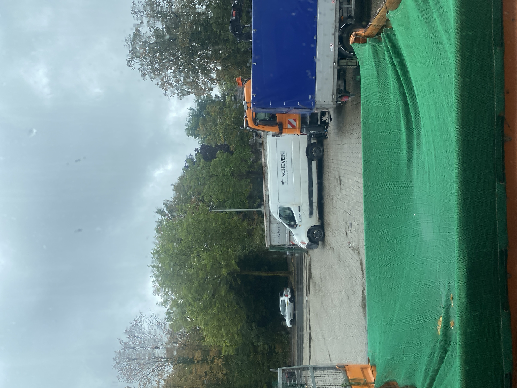 The image captures an overcast day in Erkrath, Germany, featuring a parking lot with several vehicles and a backdrop of trees. In the foreground, a bright green net is draped over an unseen object, possibly construction material or debris. To the right of the net, a grey truck with a blue tarp covering its cargo is parked. Next to it is a white delivery van with the logo "SCHEVE" visible on its side. To the left of the van, a white passenger car is parked on the side of the road, facing away from the camera. Behind the vehicles, a dense line of trees with green and autumn-colored foliage forms a natural barrier. The ground appears to be a paved surface, possibly asphalt or concrete, with some wet patches. The sky is a uniform grey, indicating cloudy weather. No people are visible in the scene.