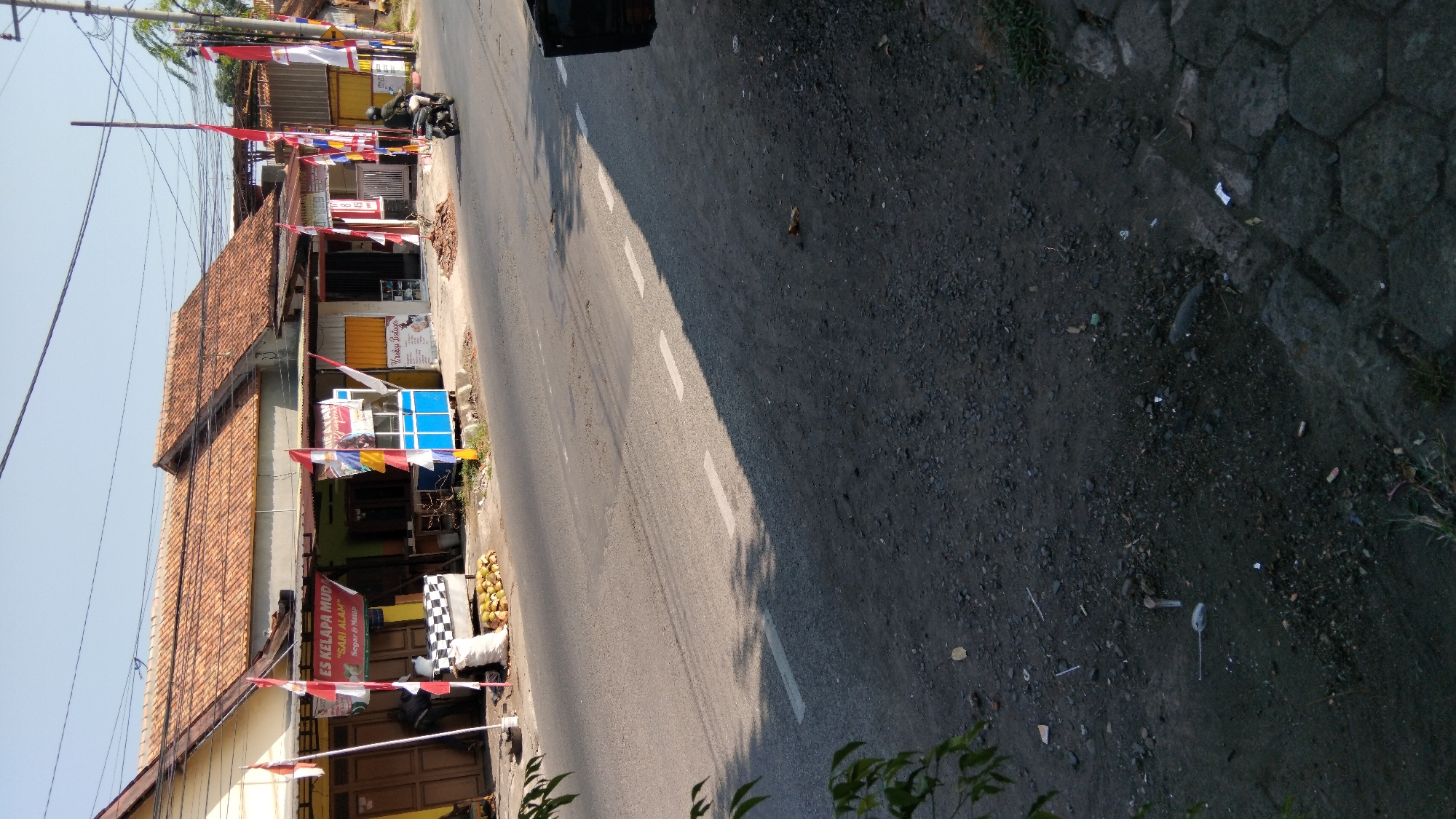 The image depicts a street scene in Proyonanggan Tengah, Indonesia, under a clear sky. In the foreground, a paved road with white dashed lines is visible, partially shaded by trees. The roadside is lined with a mixture of gravel and hexagonal paving stones, with some scattered debris.
Along the roadside, there are several small shops or stalls. One stall has a blue and white structure with a sign indicating "ES KELAPA MUDA 'SARI ALAM' Segar & Manis". Another stall displays various flags, including red and white Indonesian flags. In front of the shops, there is a pile of coconuts and a white sack. A person is seen riding a motorcycle on the road, heading away from the camera. The shops have traditional tiled roofs and their facades are painted in various colors, including yellow and white. Some of the shops have closed wooden doors and windows. Overhead, electrical wires are strung between poles. The overall impression is of a bustling roadside commercial area in a tropical climate.