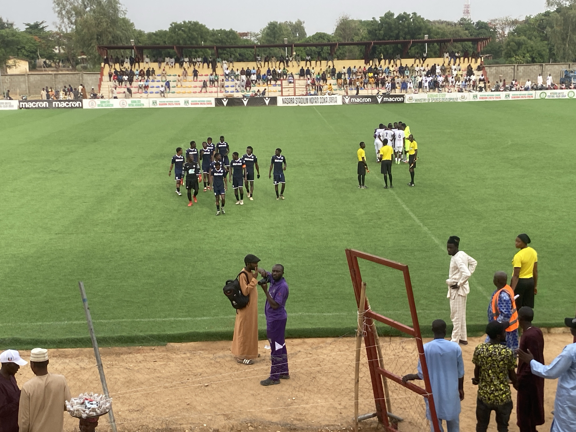 The image depicts a soccer match at Nasara Stadium Kofar Doka Zaria, in Tudun Wada, Nigeria, during the 2025/2026 season. Two teams of soccer players are on the green field, one in dark blue and white uniforms, and the other in white and black. Three referees are present, dressed in yellow jerseys and black shorts. Spectators are seated in the stadium stands behind a green advertising banner that reads "NASARA STADIUM KOFAR DOKA ZARIA". There are two men at the edge of the soccer field. One is dressed in purple soccer gear and appears to be taking a photograph. The other man is in traditional brown clothing. In the foreground are spectators standing along the perimeter fence. Some are dressed in traditional clothing, while others wear casual attire. The terrain is a dirt ground.