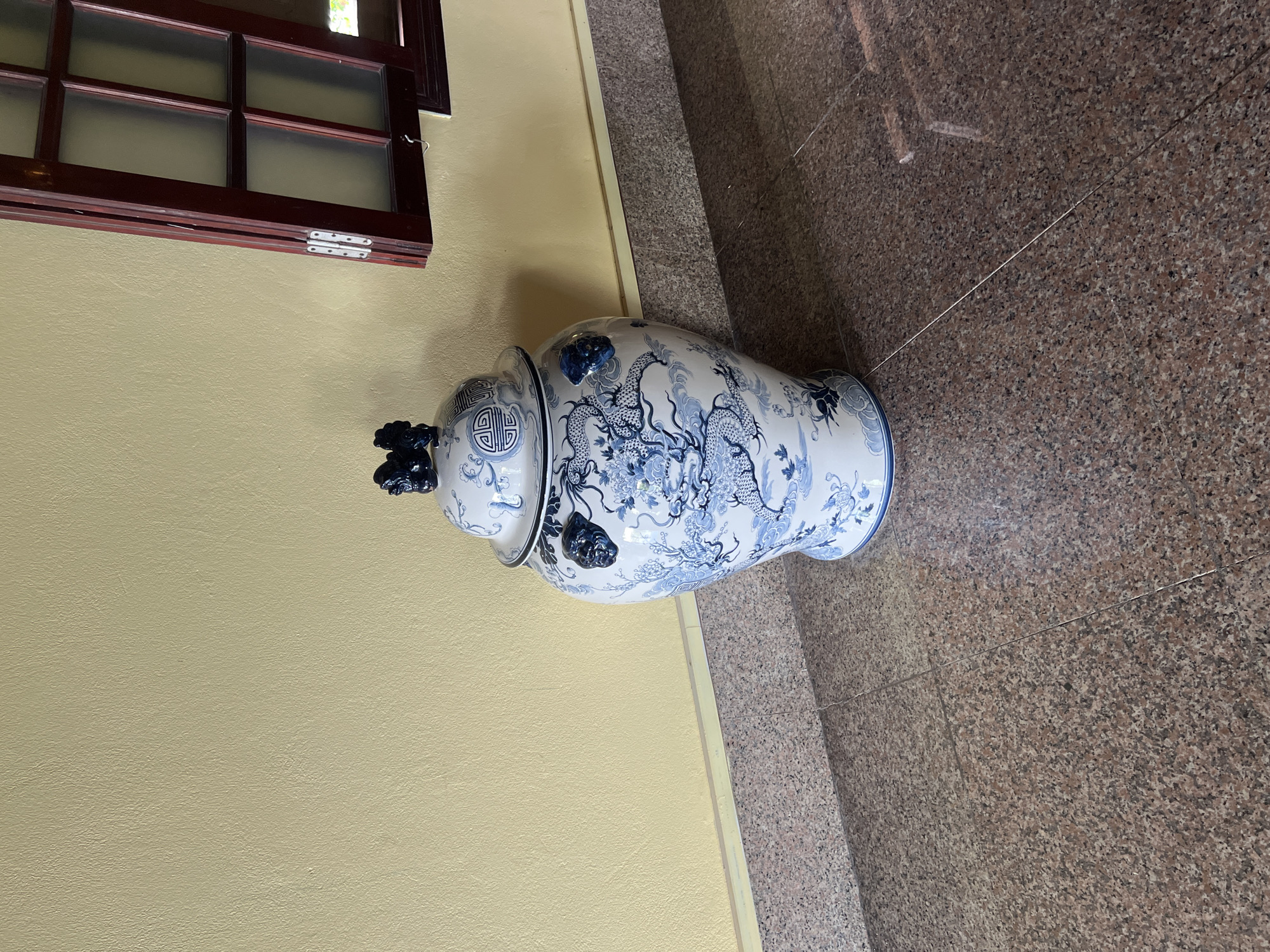 A large, ornate porcelain jar with blue dragon motifs and a lid is displayed in an interior hallway with a yellow-painted textured wall and a granite tile floor. A section of a dark wood-framed window with frosted glass is visible on the right side of the image. The jar is positioned in the center-left of the frame.