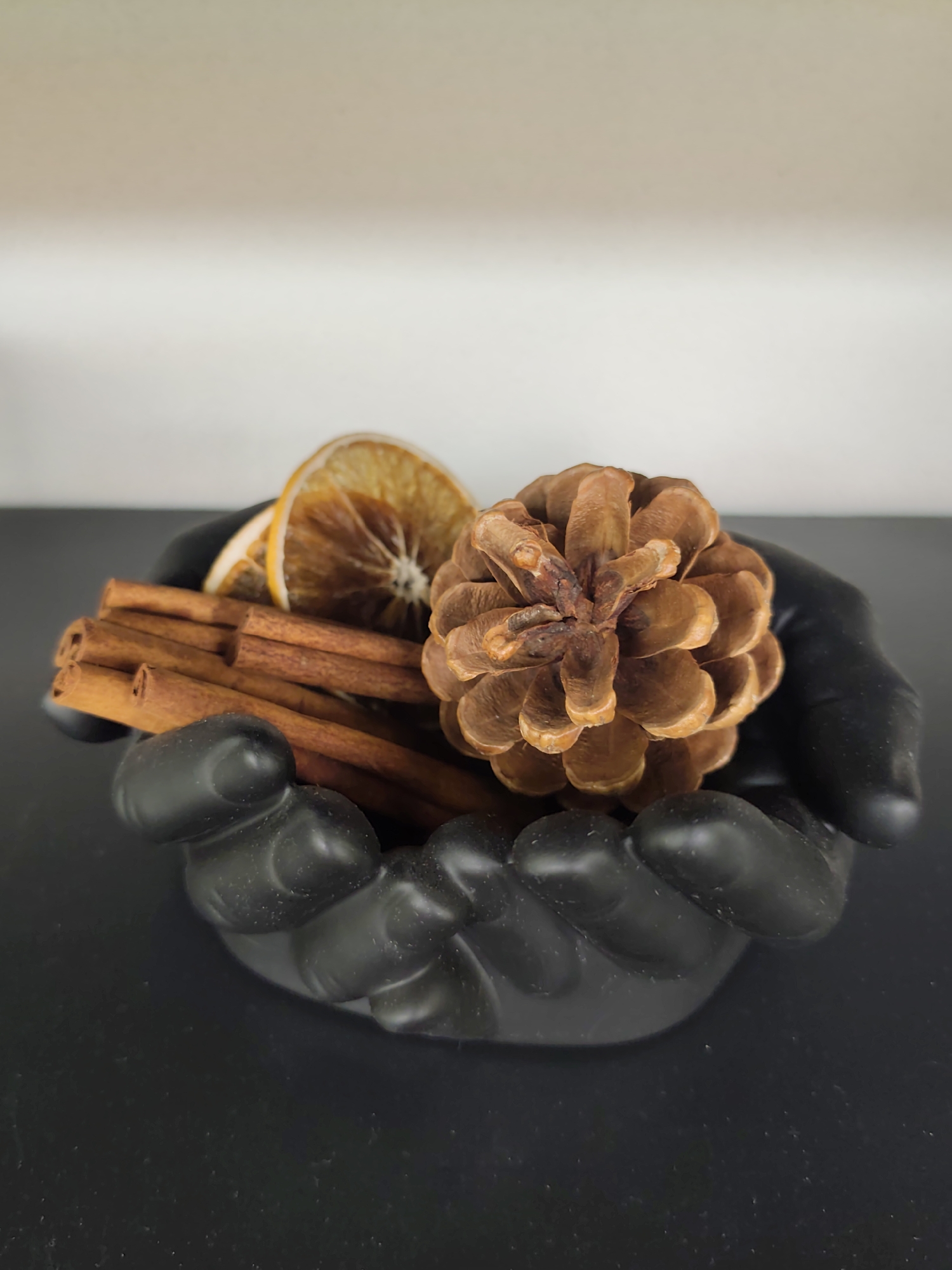 A black, hand-shaped ceramic bowl holds a collection of aromatic items on a dark, flat surface. The bowl's design mimics cupped hands, complete with sculpted fingers. Within the bowl, a dried orange slice, several cinnamon sticks, and a pinecone are artfully arranged. The background is a plain, light-colored wall. The scene is illuminated by soft, overhead lighting. No specific location in Sochi, Russia, is discernible from the image.