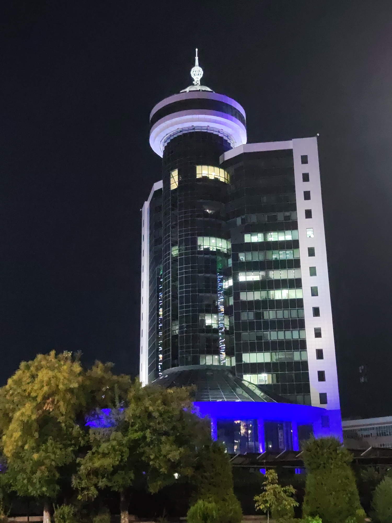 The image shows a nighttime view of the "Uzbekistan Hotel" in Tashkent, Uzbekistan. The hotel is a tall, modern building with a distinctive cylindrical top section topped with a spire. The building is illuminated, with blue lights visible at the base and white and purple lights on the upper sections. The windows of the building are lit, suggesting occupancy. In the foreground, there are several trees with green and yellow foliage, some of which are casting shadows. The overall scene depicts a well-lit urban landscape at night.
