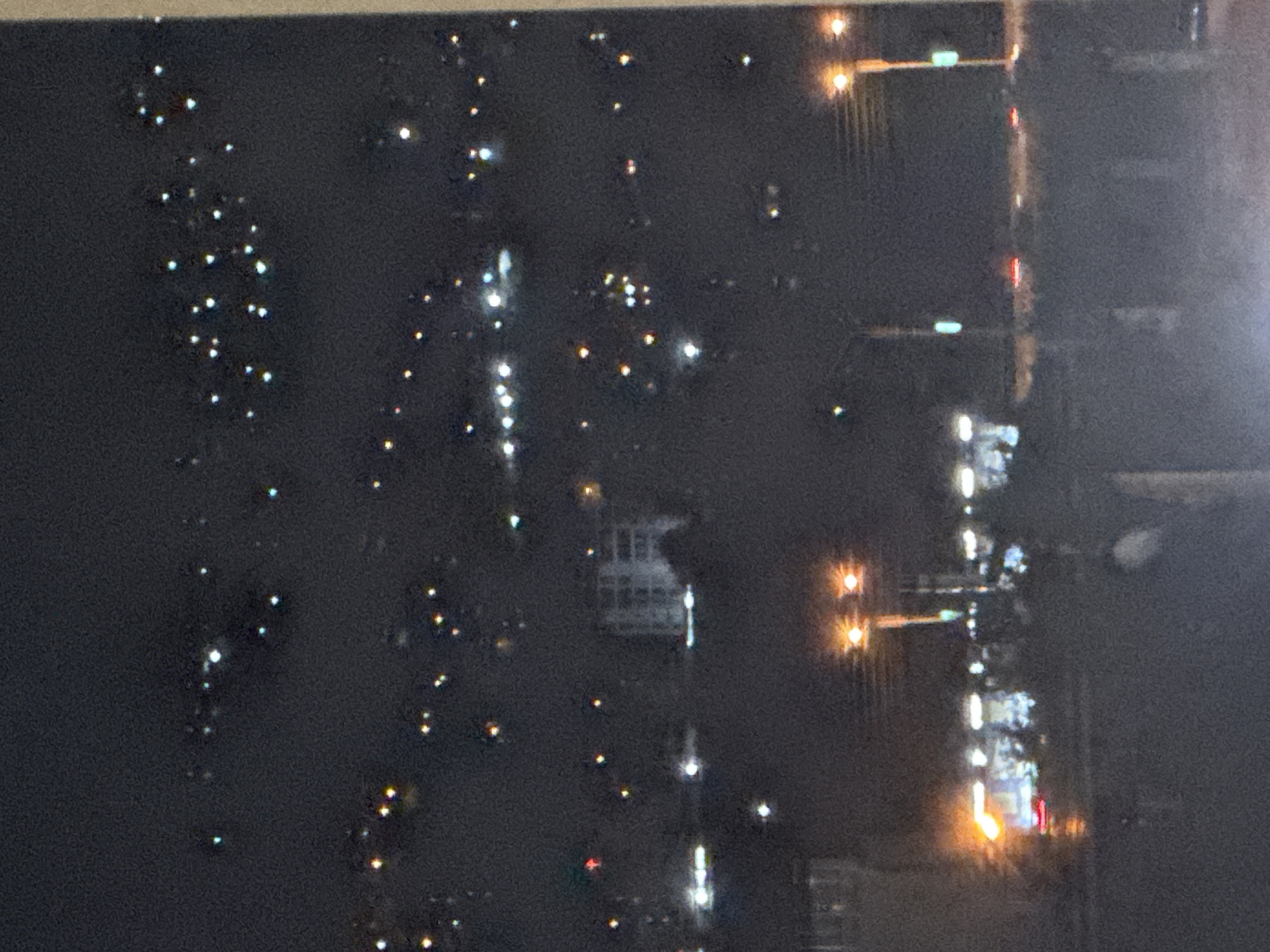 This is a night-time, slightly blurry, high-angle shot of an urban landscape, likely taken from an elevated position, such as a hillside overlooking the city. The scene depicts the spread of city lights across a dark, hilly terrain, characteristic of Tbilisi, Georgia.
In the foreground, the bottom of the image shows a blurred area, possibly a road or a balcony railing, with some indistinct shadows of people or objects. Further back, a series of bright streetlights illuminate the foreground street level, casting a warm glow. There appear to be traffic lights and possibly some businesses or residences with illuminated windows along this street.
The middle ground is dominated by the lights of buildings and structures scattered across the hills. A prominent, boxy building with multiple illuminated windows stands out amidst the darkness. Numerous other smaller lights, varying in color (white, yellow, and some hints of blue and red), pepper the slopes, indicating residential areas or smaller commercial establishments.
The background is a dense collection of city lights stretching across the horizon, suggesting a significant urban area. The lights appear to form clusters and lines, delineating the topography of the city. The overall impression is of a sprawling city at night, with a vast network of lights against a dark sky. The slight blur suggests movement or a longer exposure time. There are no distinct, clearly identifiable people or vehicles in the image, only suggestions of their presence through the ambient lighting.