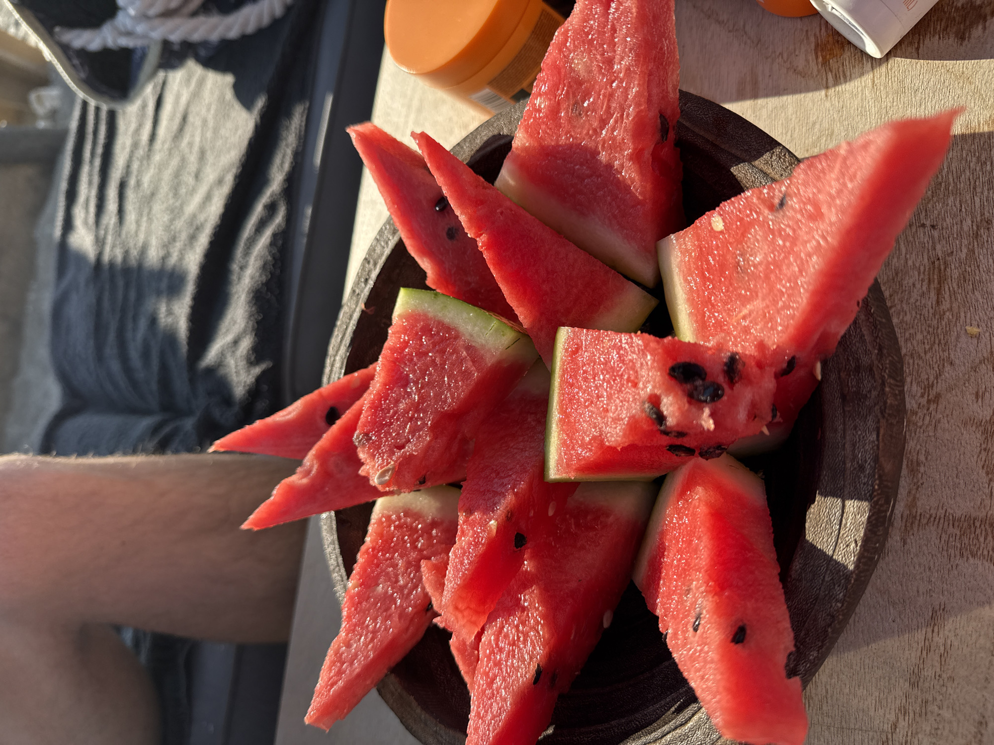 A close-up, eye-level shot shows a dark wooden bowl filled with triangular slices of ripe watermelon. The watermelon pieces are vibrant red with visible black seeds and some white rind. The bowl sits on a wooden surface, likely a table, and is partially in shadow from the sun. In the background, out of focus, a person's legs are visible, clad in dark fabric. To the right of the bowl, an orange jar, possibly sunscreen, and a small white container are also out of focus. The scene appears to be outdoors, given the strong sunlight casting shadows and illuminating the watermelon. The context of Pallini Municipal Unit, Greece is not visually discernible from the image itself.