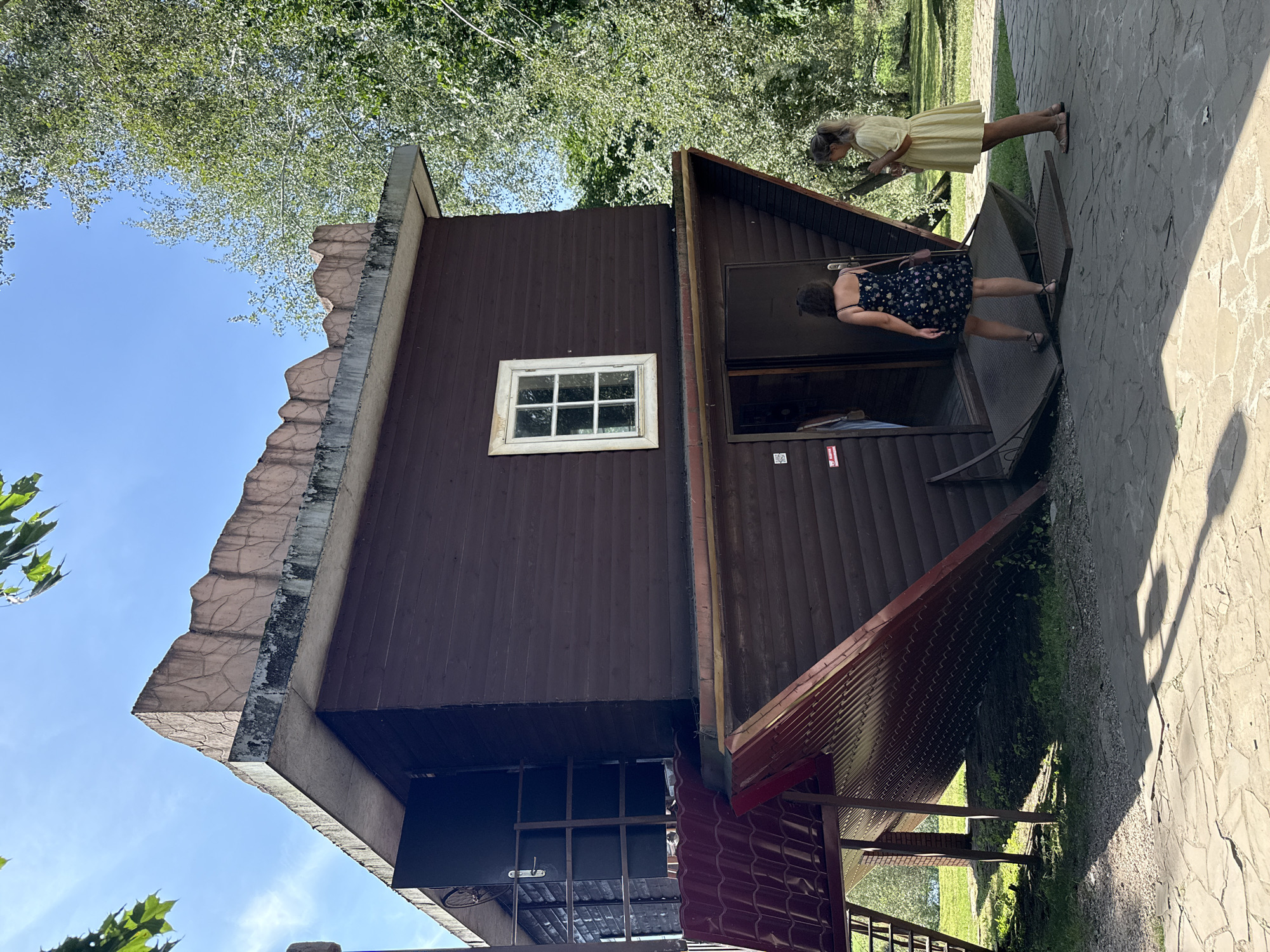 In the city of Дукорскі сельскі Савет, Belarus, an upside-down wooden house is the focal point of this outdoor scene. The house, painted a dark brown, is situated on a paved walkway, with green grass and trees forming the surrounding environment. The roof of the house, which appears to be its foundation in this inverted state, is tilted downwards, revealing a red, tiled surface. A small window with white panes is visible on the side of the house.
Two women are present in the image. One woman, wearing a dark floral print dress and sandals, is walking towards the entrance of the house. She has dark, curly hair and is carrying a small pink purse. The other woman, wearing a short yellow dress and sandals, is standing near the entrance, holding a mobile phone. Her blonde hair is pulled back.
The entrance to the house is an opening in the side, with a metal ramp leading up to it. A dark-colored door is ajar, revealing a glimpse of the interior. Signs are affixed to the wall near the entrance. The overall impression is that of a tourist attraction or an unusual architectural exhibit. The sky above is clear blue, and sunlight casts shadows on the ground.