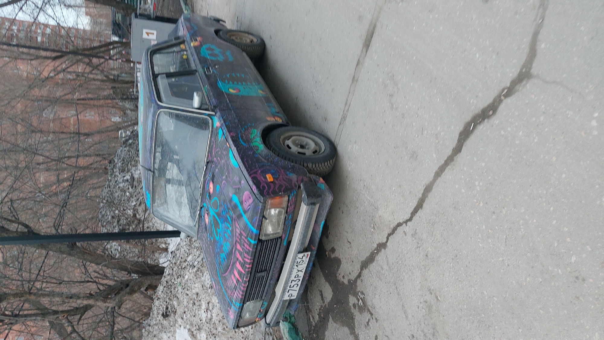 The image shows a dark-colored car with graffiti parked on a paved street, possibly in Novosibirsk, Russia. The license plate reads "P753PX 154," which indicates registration in that region. The environment includes bare trees, a lamppost, and a building in the background, suggesting an urban setting during a cold season. There are no visible people or actions taking place. The street has a visible crack running across its surface.