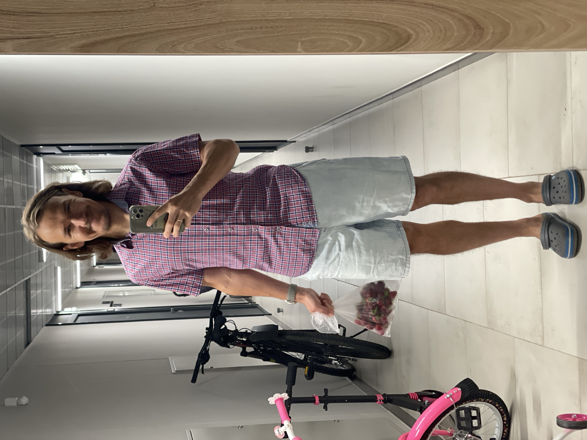 A man stands in a hallway and takes a mirror selfie. He is wearing a red and blue plaid short-sleeved shirt, light blue denim shorts, and dark gray crocs. He is holding a clear plastic bag filled with strawberries in his right hand. To his left, a black bicycle and a pink scooter are parked. The hallway has white tiled floors and walls.