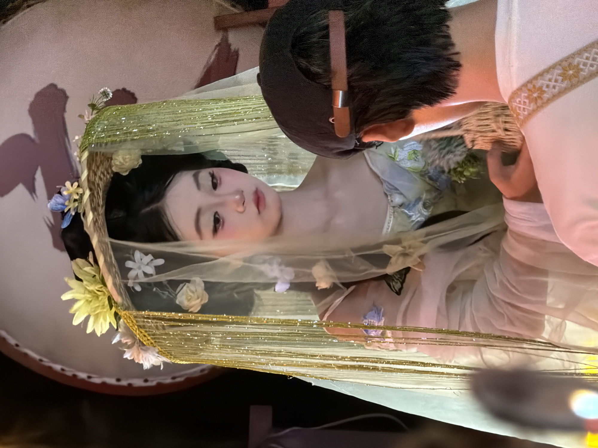 A person wearing a veiled headpiece adorned with flowers and gold fringe is looking down. A second person, seen from behind wearing a black baseball cap, is interacting with the first. The scene appears to be indoors with a drum-like object and a wall in the background. The context of Yuzhong District, China, is not visually discernible from the image itself.