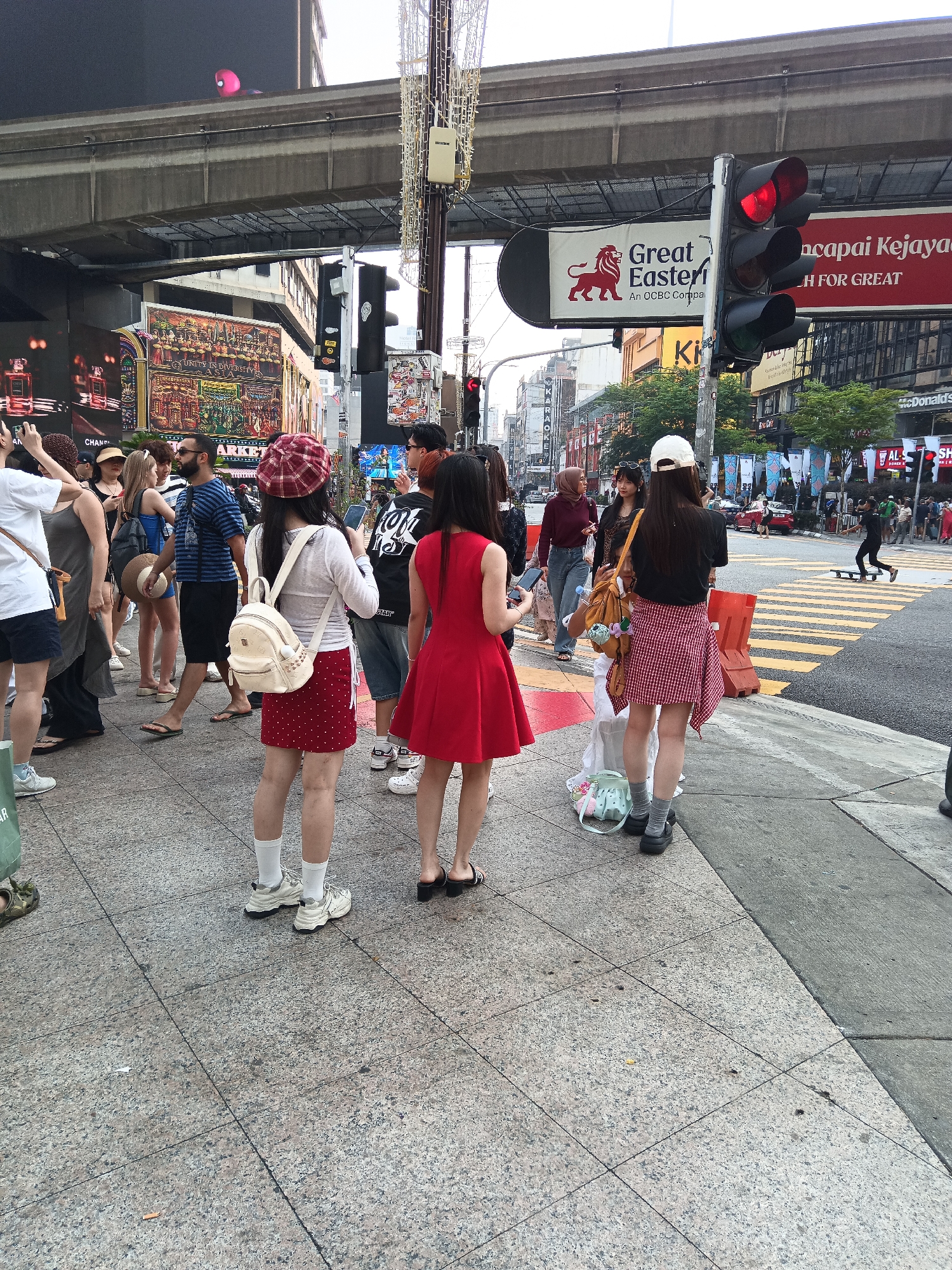 The image depicts a street scene in Kuala Lumpur, Malaysia, likely during the daytime. In the foreground, a group of pedestrians is visible on a sidewalk. One woman, facing away from the camera, wears a red and white checkered skirt, a white top, and a white baseball cap. She is holding a smartphone. To her left, another woman in a red dress stands facing away, also holding a smartphone. Further to the left, a group of individuals are gathered, some looking at their phones. In the background, a busy street with cars and a skateboarder crossing a pedestrian lane is visible. Traffic lights are red, indicating a stop. Buildings line the street, featuring various advertisements and signage. An elevated train track runs across the top of the image. The overall environment suggests an urban setting with considerable pedestrian and vehicular traffic.