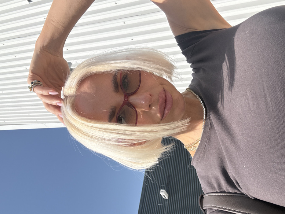A female subject with platinum blonde, bob-cut hair, featuring subtle pink streaks on the left side, is depicted in an outdoor, chest-up, slightly low-angle shot. She wears dark red-framed tinted sunglasses, a dark gray short-sleeved t-shirt, a thin silver-toned choker, and a thicker gold-toned necklace. Her right hand, adorned with light pink manicured nails and multiple rings, is positioned with fingers resting on the crown of her head. A dark gray bag strap is visible over her left shoulder. The background consists of a clear blue sky above two distinct industrial-style structures: a dark gray horizontally corrugated building with a white rectangular fixture on the left, and a brightly illuminated white vertically corrugated building on the right. The scene is set under direct daylight in Dubai, United Arab Emirates.