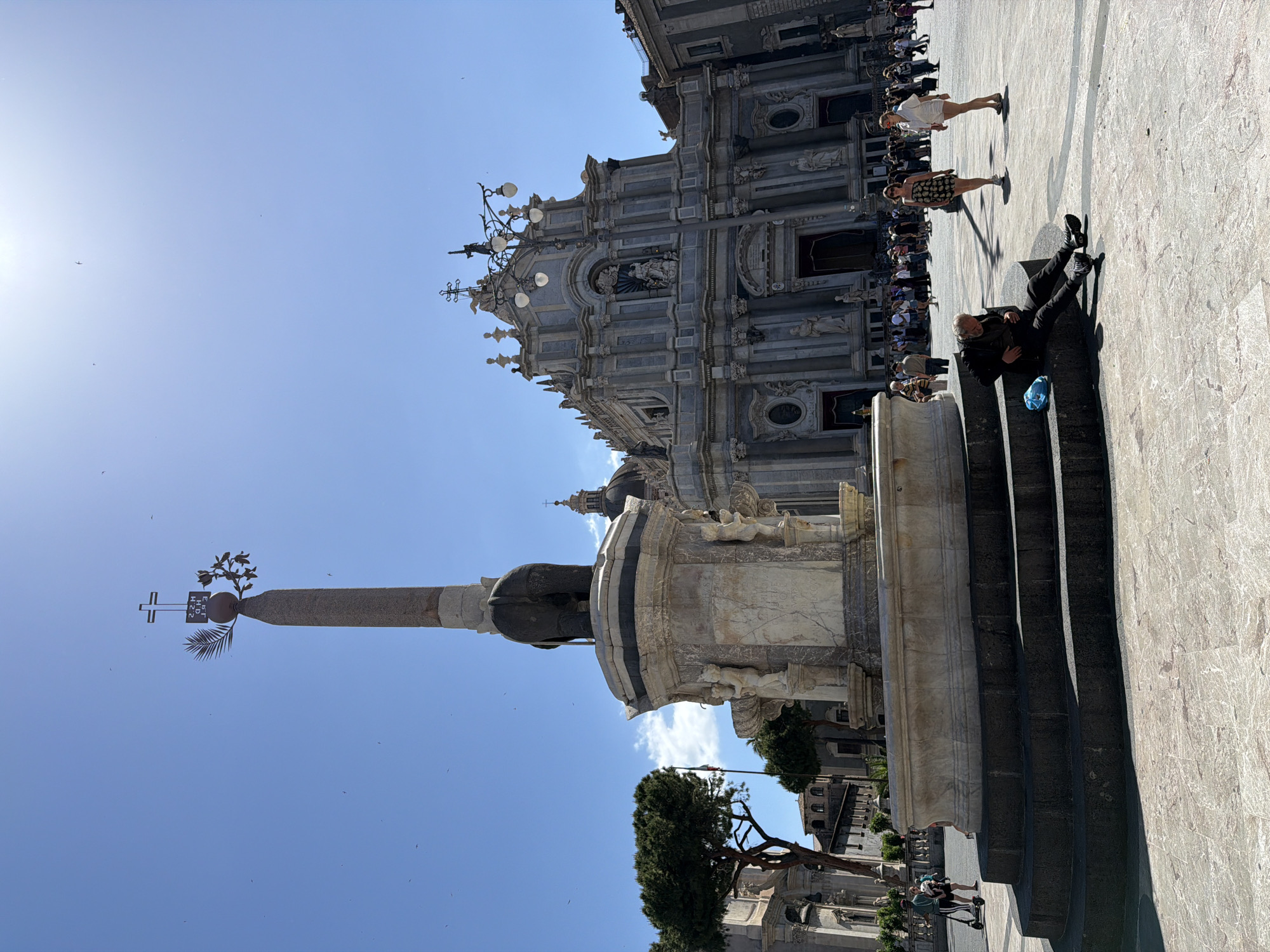This photograph captures a sunny scene in Catania, Italy, focusing on Piazza del Duomo. A prominent element is the Elephant Fountain, a statue of an elephant topped with an obelisk. Behind the fountain stands the Catania Cathedral, displaying its ornate baroque facade. People are visible in the background, some standing near the cathedral and others walking across the plaza. A man is sitting on the steps of the fountain, seemingly resting or observing the surroundings. The sky is clear and blue.