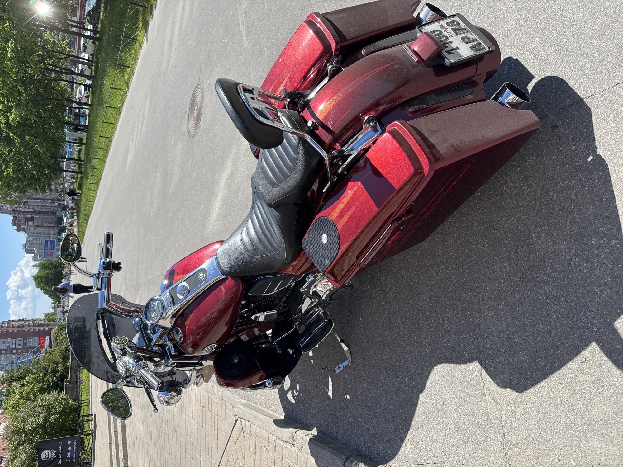This image features a glossy, deep red touring motorcycle, likely a Harley-Davidson, parked on a paved surface next to an asphalt road. The motorcycle is the central subject, showcasing its metallic paint with a sparkling finish, chrome accents on the engine, handlebars, mirrors, and exhaust tips. It is equipped with a large front fairing and windshield, a black padded solo seat, a chrome sissy bar with a luggage rack, and color-matched hard-shell saddlebags.
The rear of the motorcycle clearly displays a Russian license plate with the sequence "1100 AP 78 RUS." The "78" regional code indicates Saint Petersburg, Russia, matching the provided location context.
The scene unfolds on a bright, sunny day, evidenced by the clear blue sky with scattered white clouds and the strong, well-defined shadows cast by the motorcycle and surrounding elements. This suggests a time around mid-day. In the background, a line of green trees and a grassy area with a fence separate the parking spot from a road where several cars are visible. Urban buildings extend into the distance, and faint figures of people can be seen moving about. A manhole cover is noticeable on the paved ground. One cropped view reveals a sign for "GALAXY GAMES" that also mentions "PLAY & WIN | GAMES | CAFE," indicating a commercial establishment nearby. The overall setting is an active urban environment in Saint Petersburg, Russia.