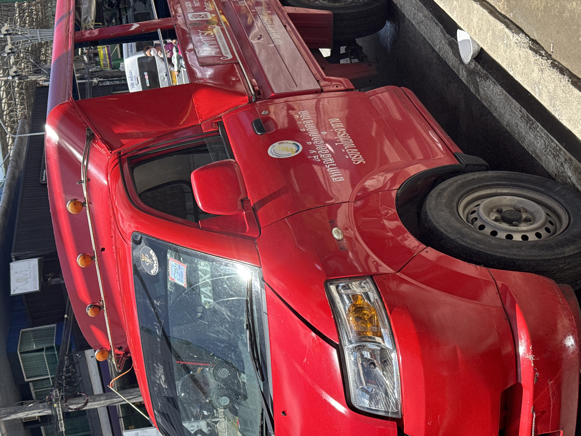 The image shows the side view of a red taxi truck parked on a street in Patong, Thailand. The truck has a passenger compartment with a canvas roof. Several logos and text are visible on the truck's exterior, including the word "TAXI" on the windshield. The surrounding environment appears to be an urban area with buildings and power lines visible in the background. The street is paved, and a curb is present in the foreground. A white plastic bowl is on the ground near the curb. A person wearing a face mask is visible inside the passenger compartment of the truck. The vehicle has visible wear and tear, with scratches and dents on the red paint.