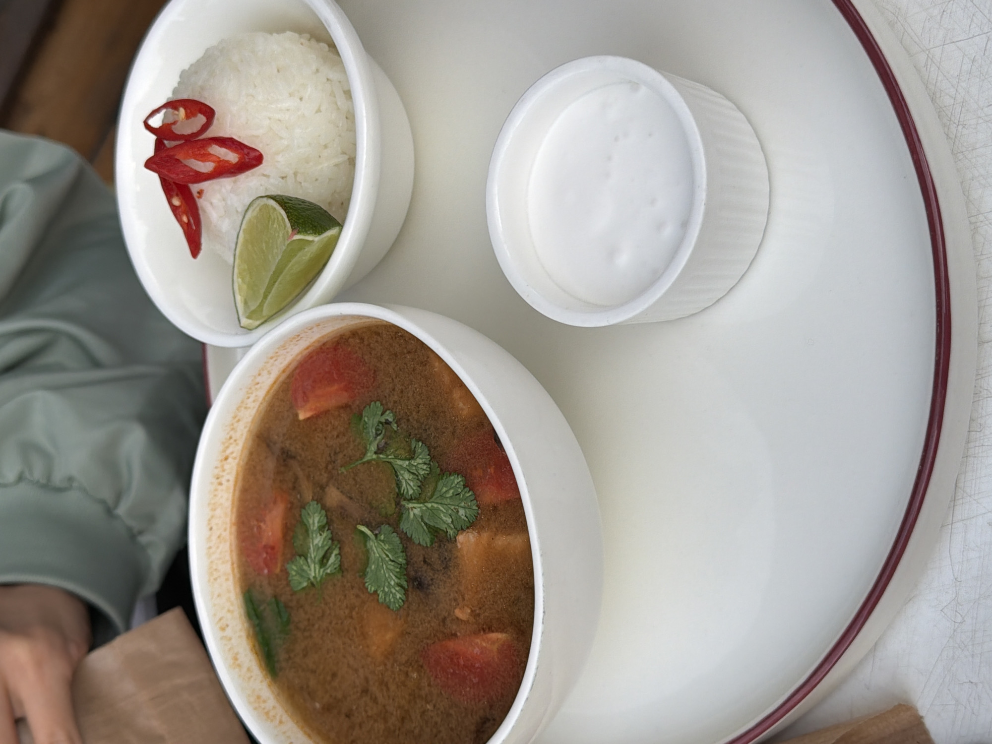 An overhead shot shows a dining setup, likely in a restaurant in Yerevan, Armenia, featuring three white bowls on a large white plate with a red rim. The bowl at the lower left contains a soup with visible tomatoes and green herbs floating within. A bowl in the upper left contains white rice topped with slices of red chili and a wedge of lime. A third bowl, positioned towards the upper center, holds a white liquid, possibly coconut milk or cream. A portion of someone's arm and green jacket are visible in the lower left of the frame. The plate sits on what appears to be a white surface.