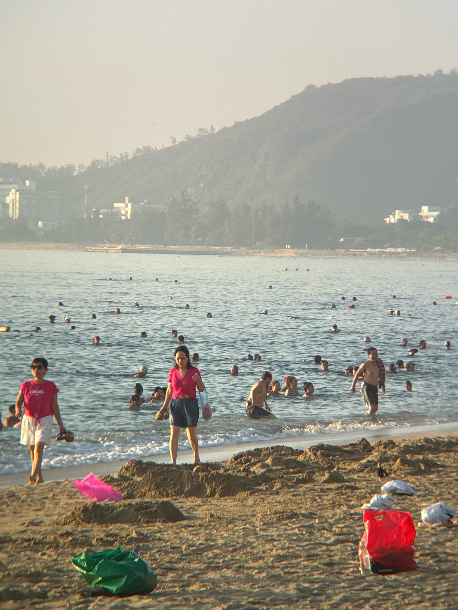 This image depicts a crowded beach scene in Nha Trang, Vietnam, likely during the daytime given the lighting. In the foreground, a sandy beach is littered with several items: a bright pink plastic sheet, a crumpled green plastic bag, and a bright red plastic bag with white items inside, possibly plastic bottles or bags. Two women are walking on the beach near the water's edge. The woman on the left wears a pink t-shirt and white shorts, holding a pair of dark sandals. The woman on the right wears a pink top and dark shorts, also holding sandals and a plastic bag.
The ocean is filled with numerous people swimming and wading. Many heads are visible above the water. Further out, the coastline is lined with buildings and lush green hills, characteristic of Nha Trang's landscape. The atmosphere appears hazy, suggesting it might be a warm or humid day, or there could be some level of air pollution. The overall scene conveys a lively beach atmosphere with many people enjoying the water.