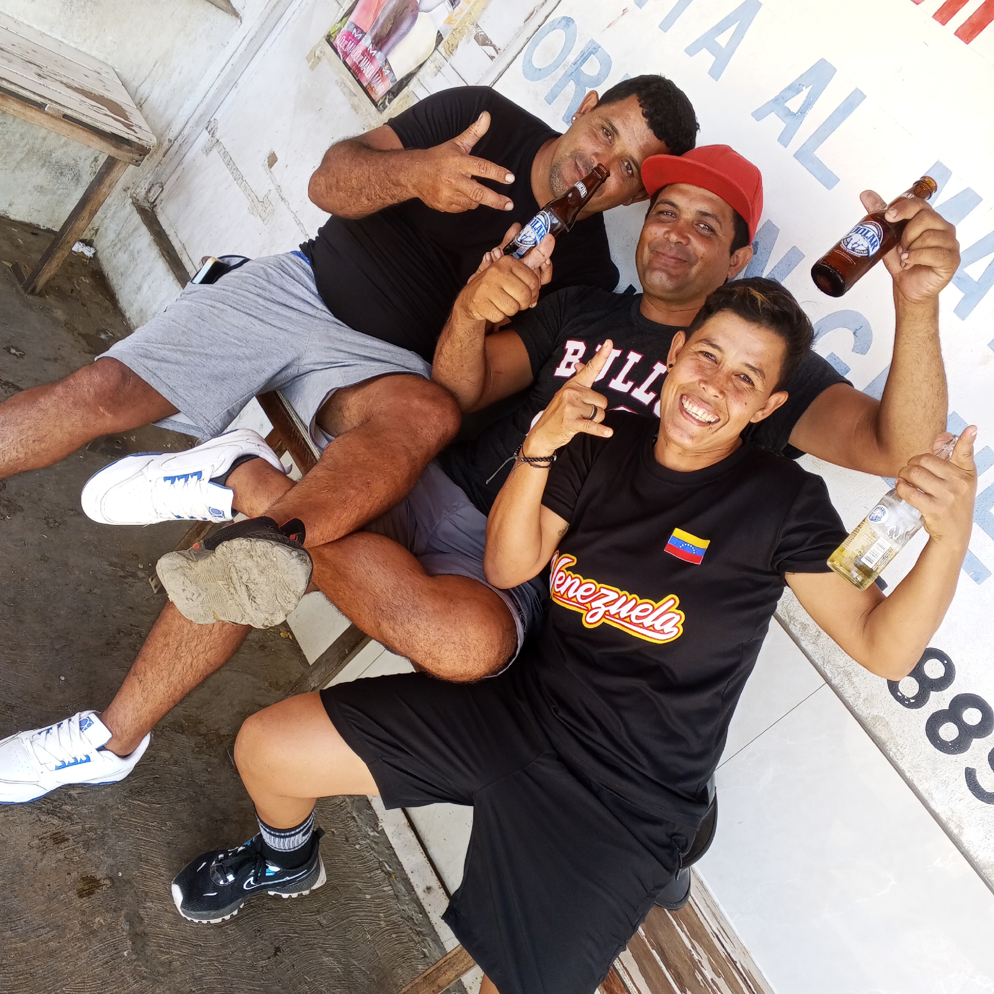Three men are captured in a vibrant, slightly tilted, high-angle full shot, seated and posing for the camera in what appears to be an outdoor, casual setting. All three subjects are smiling broadly, conveying a sense of happiness and camaraderie.
The man on the left, with dark hair, a beard, and wearing a black t-shirt and grey shorts, sits with one leg crossed over the other. He holds a brown beer bottle, identified as "Pilsener," near his face and makes a "peace" sign with his other hand. His white sneakers with blue accents are visible.
The man in the middle, wearing a red baseball cap, a black t-shirt with "BULLS" written on it, and grey shorts, also holds a brown beer bottle with "Pilsener" visible on the label. He is positioned slightly behind and to the right of the first man, looking towards the camera with a wide smile.
The third man, positioned to the right and slightly in front of the middle man, wears a black t-shirt with a Venezuelan flag patch and the word "Venezuela" in yellow and red script. He holds a clear glass beer bottle, possibly "Polar" (though the label is less clear), and gives a thumbs-up gesture. He has short dark hair and a bright smile. His black sneakers with white soles are visible on the ground.
The background consists of a light-colored, possibly white, wall with some faded text. Partially visible text includes "ITA AL MA" in blue letters and a series of numbers, "88," in black at the bottom right. A simple wooden table is seen in the upper left, suggesting a modest, outdoor area. The ground beneath the men is a mix of concrete and possibly dirt, with visible textures. The bright lighting suggests it is daytime.