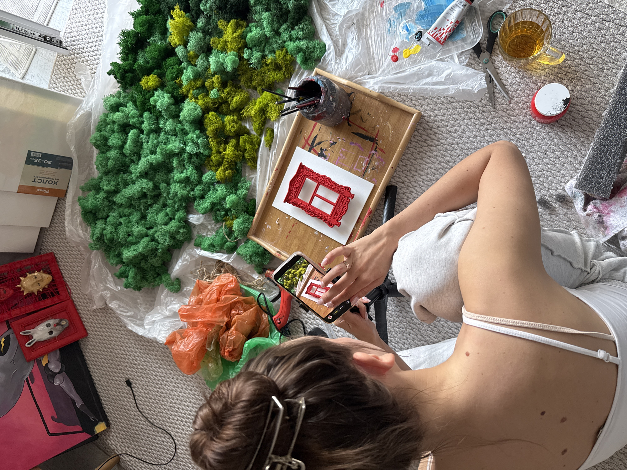 A high-angle shot shows a person sitting on a light-colored carpet, surrounded by art and craft supplies. The person, with dark hair pulled back in a bun, is taking a photo with a smartphone of a small red frame placed on a tray. The tray also holds a jar filled with paintbrushes.
Around the person, various art-related items are scattered: a pile of green and yellow moss, orange plastic bags, paints, scissors, and a mug filled with liquid. A red plastic container with small animal figurines is also visible. A painting with red, gray, and black colors rests at the lower-left corner of the image.
The environment appears to be an indoor space, likely a home studio or craft area. There is no definitive evidence of the location being Krasnoyarsk, Russia.