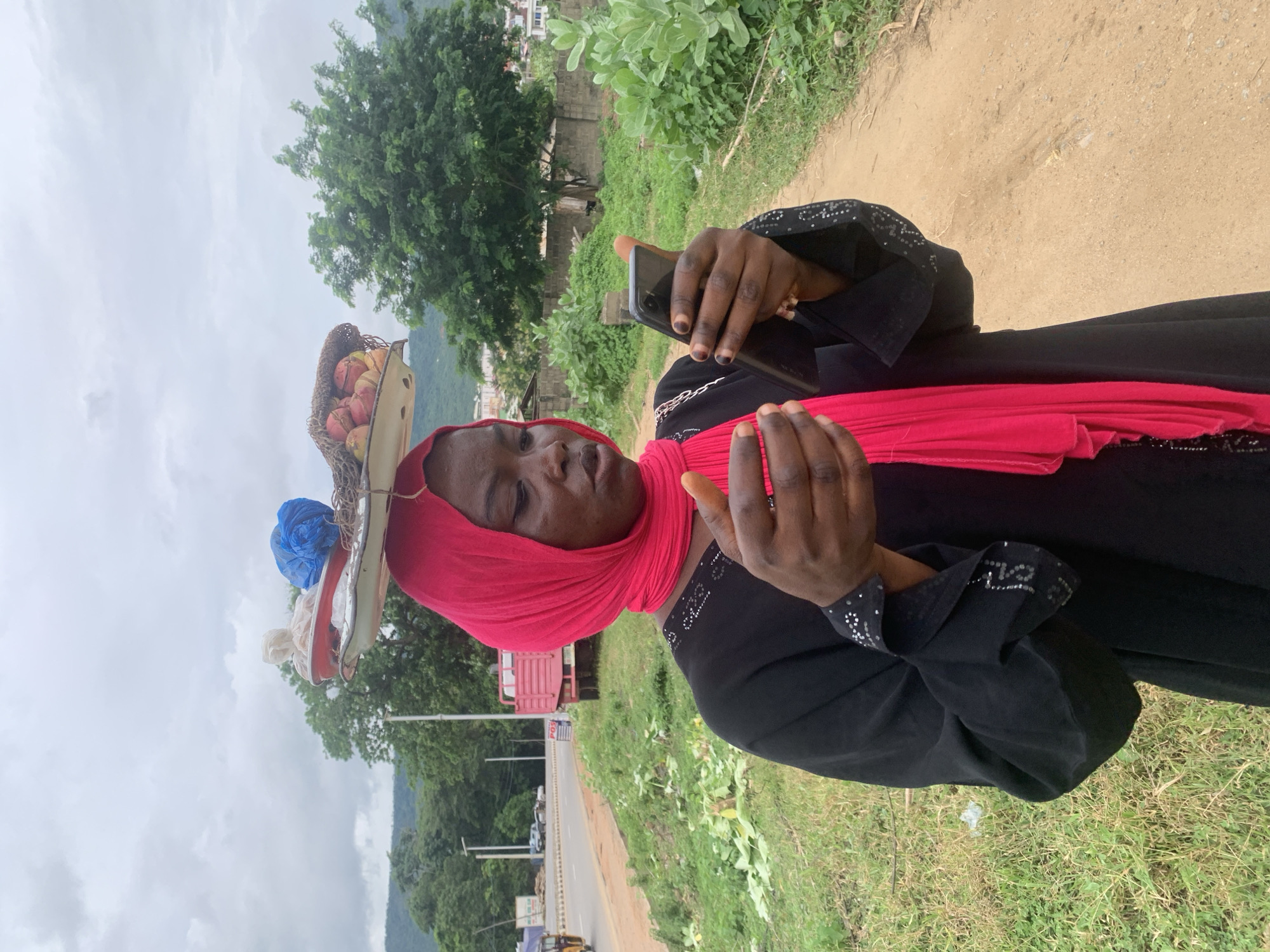 A woman, identified as being in Adankolo, Nigeria, stands on a dirt path next to a grassy area and a road. She wears a black abaya with embellished sleeves and a vibrant pink hijab. A large platter is balanced on her head, containing various items including what appear to be fruits, a blue plastic bag, and white material. She is looking down at a black smartphone held in her hands, seemingly engrossed in its use. In the background, there is a road with vehicles, trees, and buildings, suggesting a semi-urban environment. The sky is overcast.