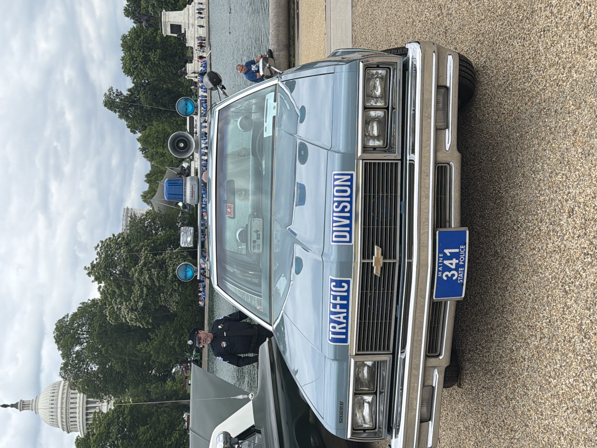 The image shows the front of a light blue vintage police car with "TRAFFIC DIVISION" written on the hood. The car has a Maine State Police license plate, number 341. The US Capitol building is visible in the background, indicating the location is in Washington, D.C. A body of water is adjacent to the car and the Capitol building. People are standing along the edge of the water. A police officer in uniform stands near the car. There are various light and speaker attachments on the car. The ground is a gravel surface.