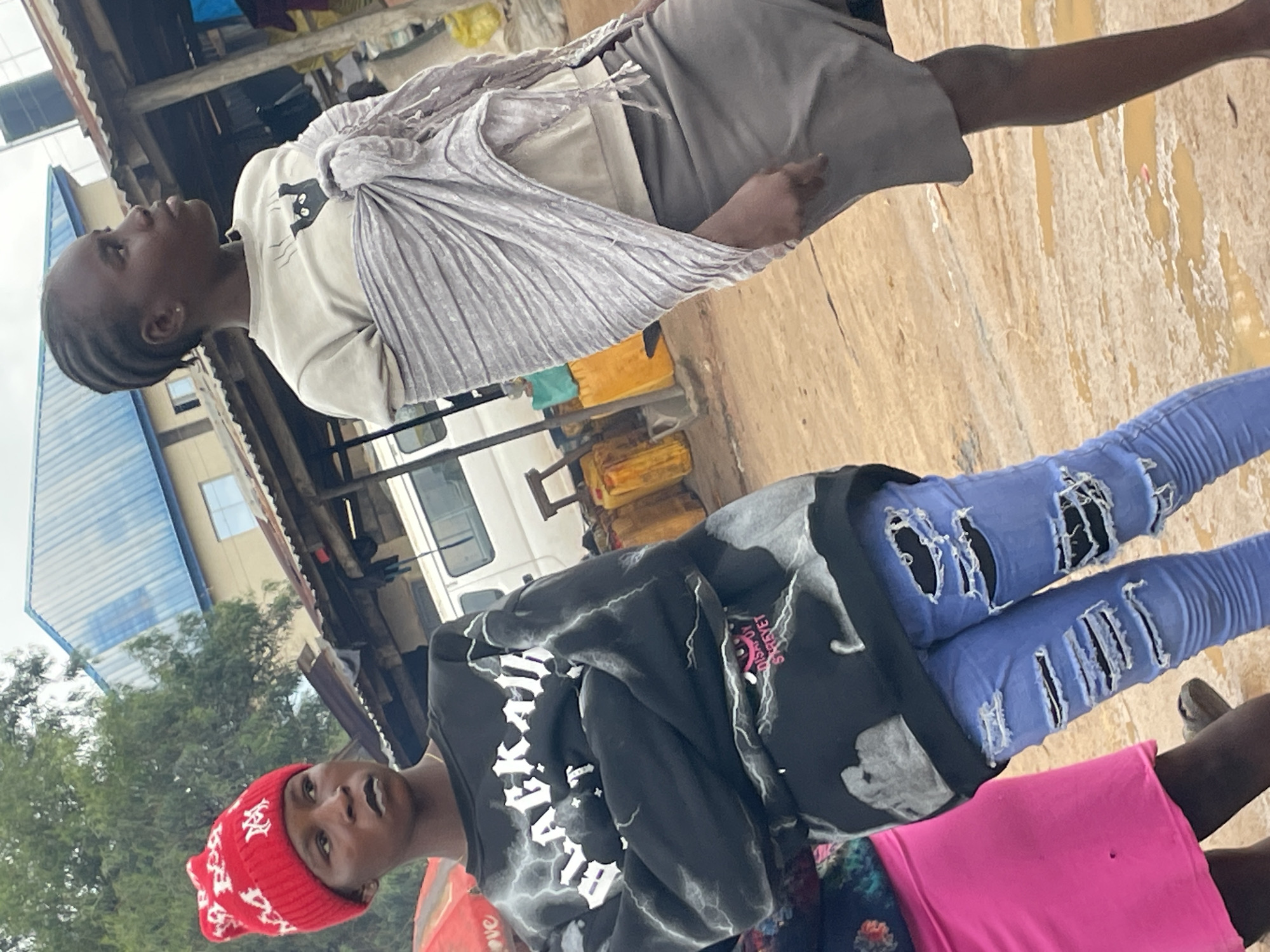 The image depicts two young women in an outdoor marketplace in Wang, Nigeria. The woman on the left wears a red beanie with white lettering, a black sweatshirt with white lightning bolt designs and text, and ripped light blue jeans. She is looking towards the left of the frame with her arms crossed. The woman on the right wears a light-colored t-shirt with a black cat graphic and a gray skirt. She is wrapped in a striped gray shawl and is looking to the right. Behind them, various market stalls are visible, including a white van and several yellow jerrycans. The ground is dirt and appears wet in places, with puddles of water reflecting the surroundings. The overall impression is a bustling, everyday scene in a Nigerian market.