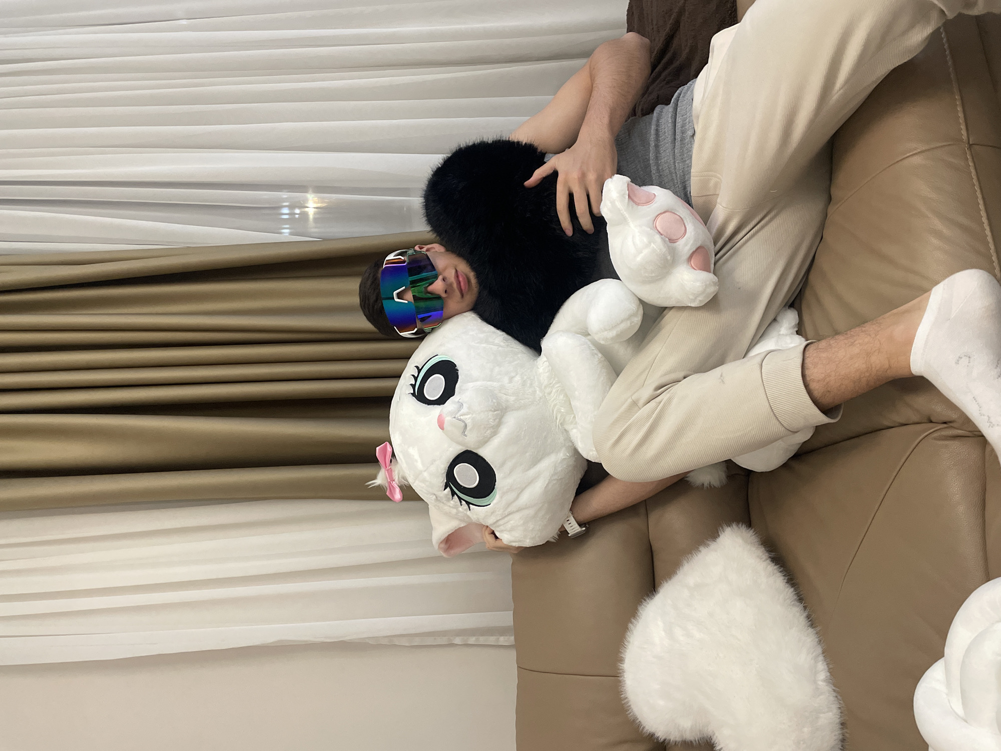 A person wearing iridescent ski goggles is reclining on a brown leather couch. The individual is wearing beige pants and a grey shirt, with one white sock visible on their right foot. They are surrounded by several plush objects: a large white cat-shaped stuffed animal with a pink bow, a black furry object, and a white paw with pink spots. A white, heart-shaped fluffy object is also visible on the couch. The background consists of white and brown draped curtains. The location is not discernible from the image.