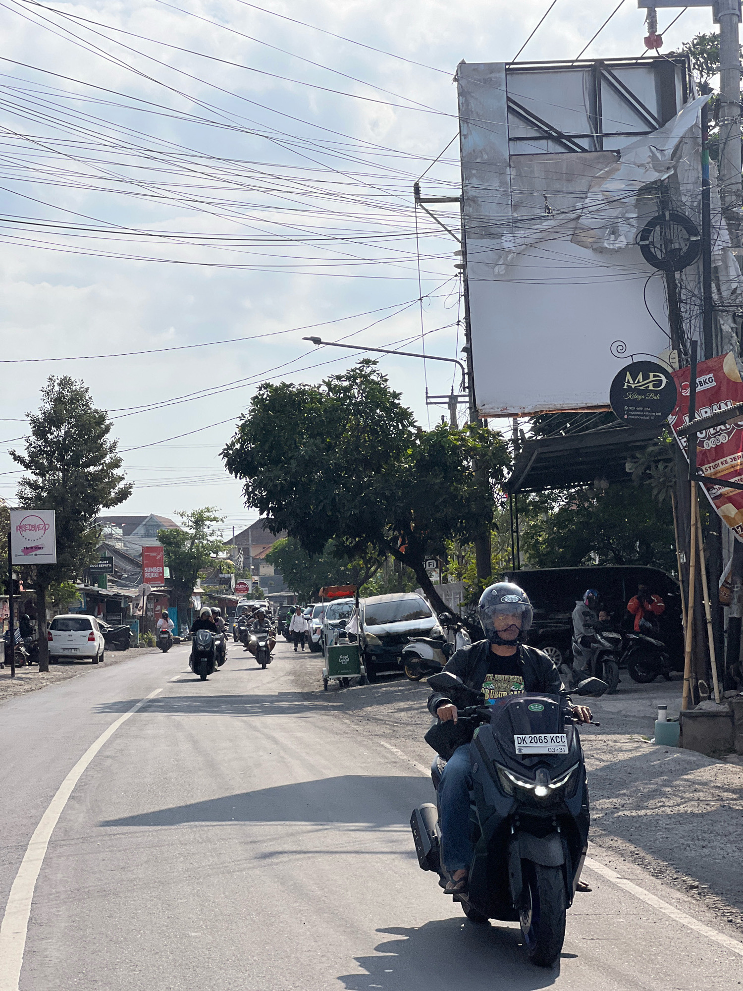The image depicts a street scene in Denpasar, Indonesia, with a focus on a motorcycle rider in the foreground. The rider, wearing a helmet and a dark jacket over a graphic t-shirt and jeans, is on a modern, dark-colored scooter with the license plate "DK 2065 KCC". The scooter has its headlights on.
The street is paved with asphalt and marked with a white line on the left. The scene is bustling with traffic; several other motorcycles are visible on the road, some with riders and others parked. Cars, including a white sedan and a dark van, are also present. The environment is characterized by a mix of urban elements: buildings line the street, some appearing to be shops with signage. Trees with lush green foliage are scattered along the roadside. Overhead, a complex network of electrical wires crisscrosses the sky against a bright, partly cloudy backdrop. A large, partially covered billboard is prominently positioned on the right side of the image, accompanied by numerous utility poles and wires. The overall lighting suggests it is daytime.