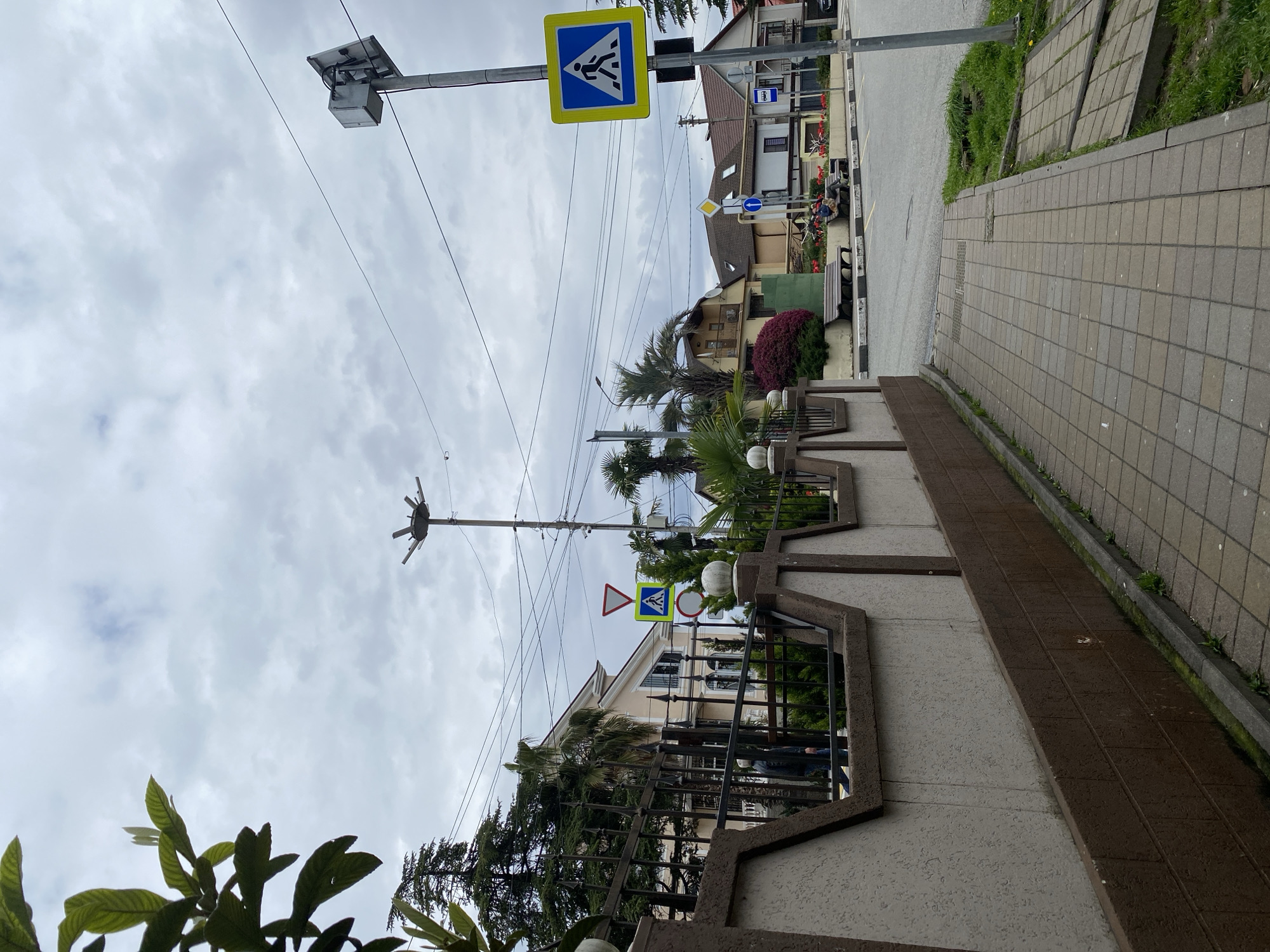 The image shows a street scene in Sochi, Russia, characterized by cloudy skies and a combination of residential and infrastructural elements.
**Environment**: The scene unfolds along a pedestrian walkway, marked by paving stones and bordering a road. The sky is overcast. Vegetation, including palm trees and other greenery, is prominent throughout the scene.
**Objects**: Several elements stand out in the scene, including power lines crisscrossing above the street, street lamps, and traffic signs, including pedestrian crossing signs. A segment of a fence runs parallel to the sidewalk, delineating private property.
**Actions/Interactions**: There is minimal visible activity in the scene. A couple of indistinct figures can be seen in the background.
**Subjects**: The main subjects of the image are the built environment and street infrastructure. The focus is on the arrangement of elements in an urban setting.