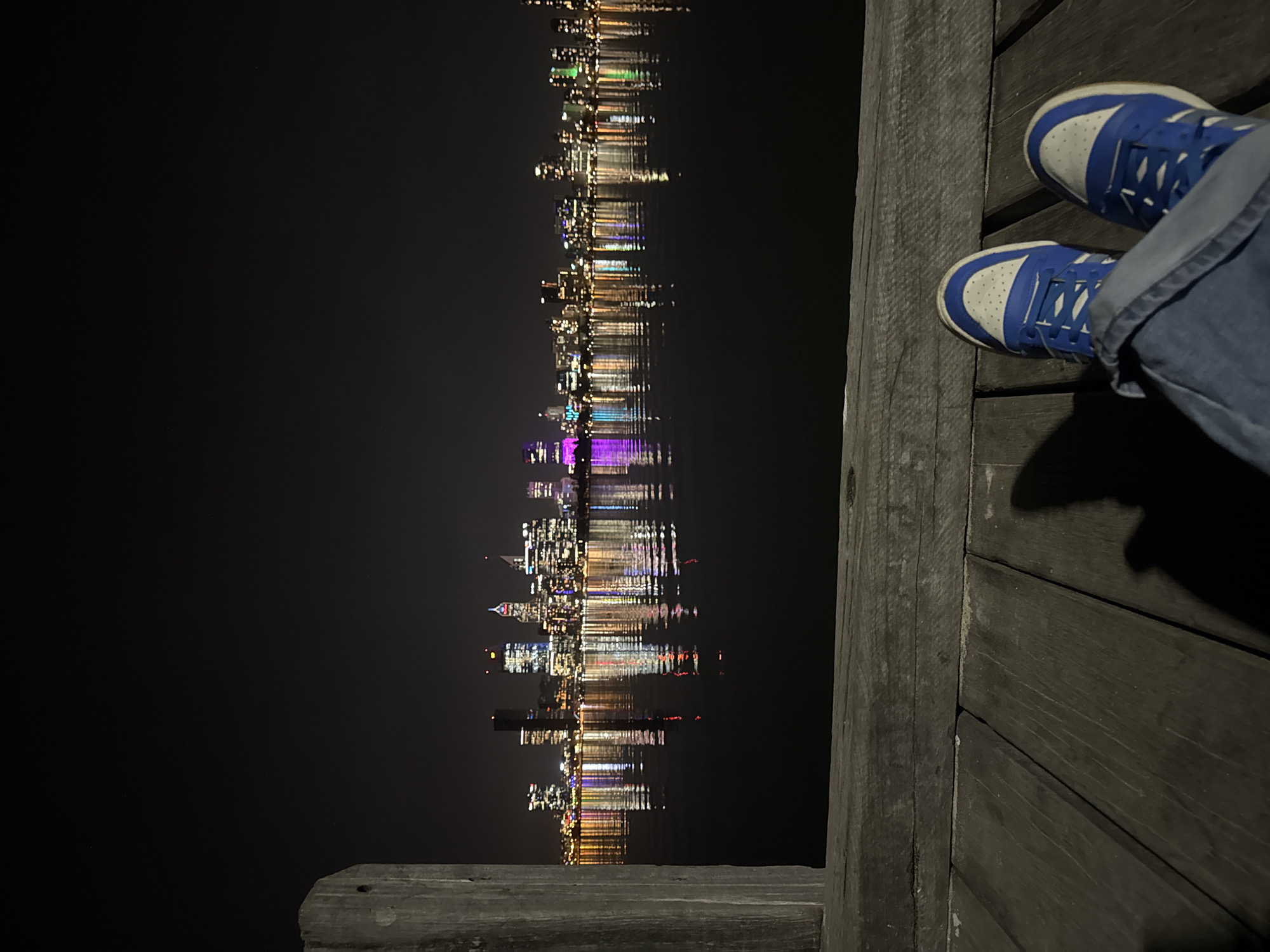 At night, the image shows a person wearing jeans and blue and white sneakers sitting on a wooden pier in South Perth, Australia, overlooking the Swan River. The city skyline is reflected in the water. The person's shadow is cast on the wooden boards.
