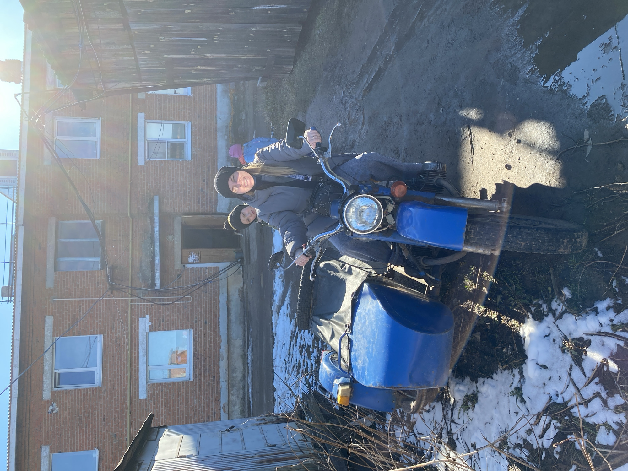 Two individuals are positioned on a blue motorcycle with a sidecar. The setting appears to be in a rural or semi-rural area, possibly on the outskirts of Noviy, Russia, indicated by the presence of older brick buildings and a muddy, snow-patched ground. The woman on the motorcycle has a visible smile. The photograph is taken during the daytime.